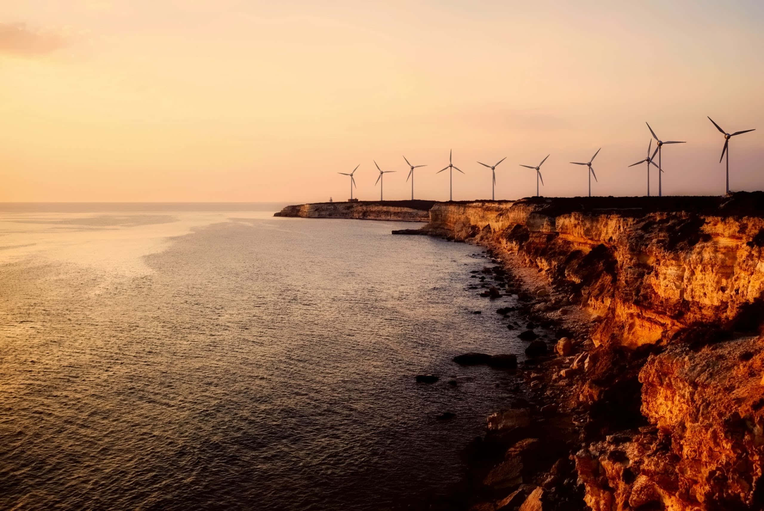 Wind 桌布 壁紙 — 風力發電機在崎嶇的懸崖上形成剪影，金色的夕陽光芒以溫暖的色調沐浴著海洋和岩石。
