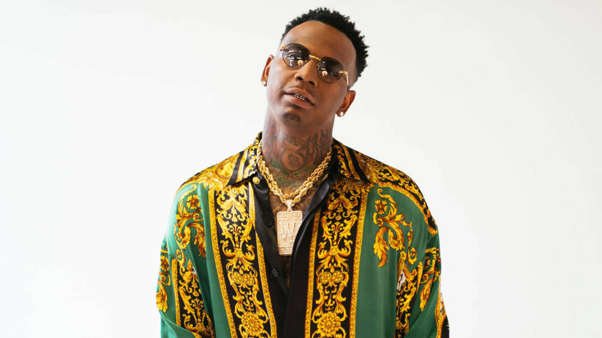 風度翩翩 Moneybagg Yo 壁紙 — 一位時尚男士，身上有紋身和戴金鏈，穿著綠色和金色圖案襯衫，背景為純白色。