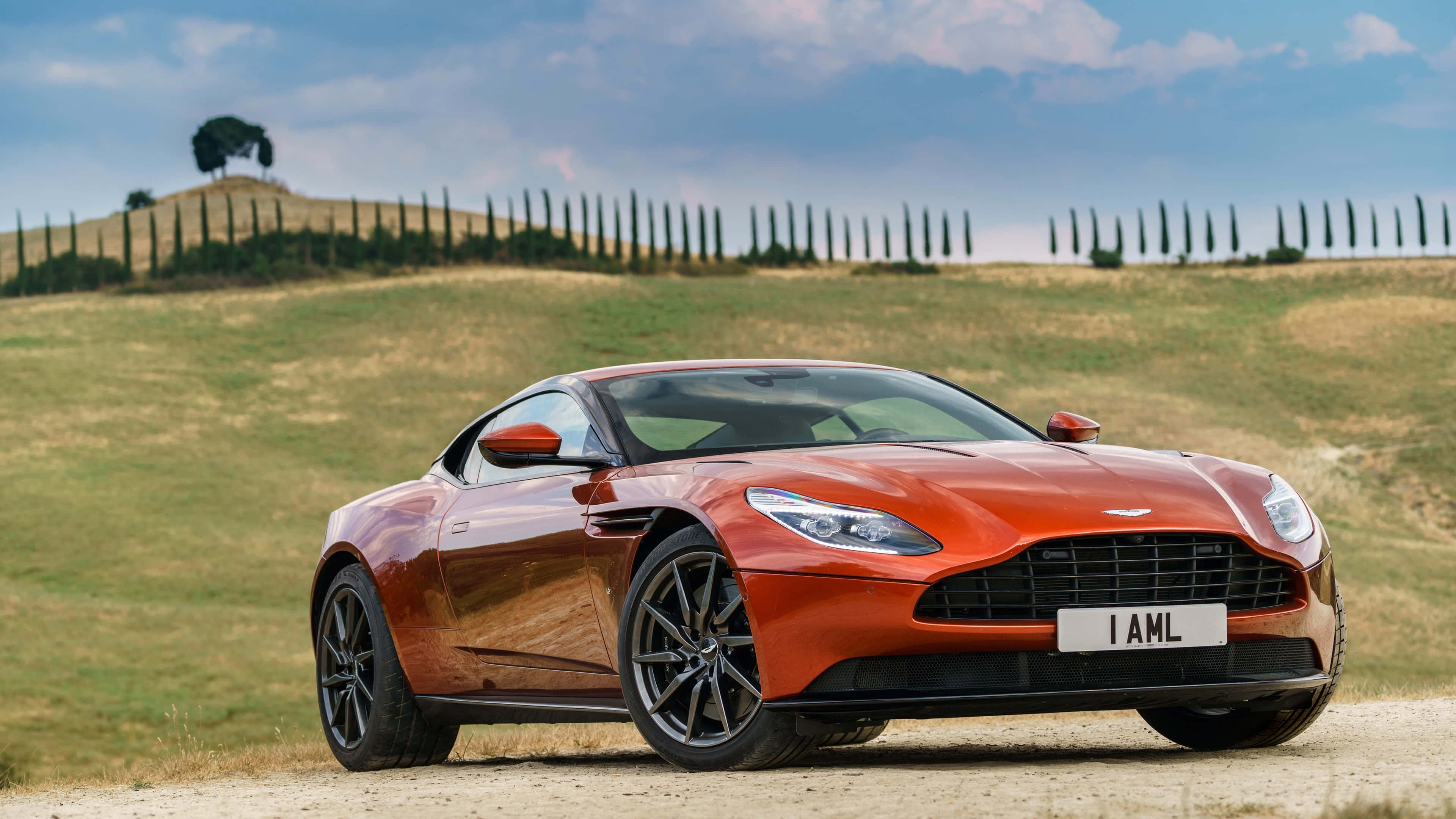 風格、速度、奢華 — 阿斯頓·馬丁 DB11 壁紙 — 一輛時尚的橙色Aston Martin跑車停泊在托斯卡納起伏山丘中的一條泥路上，周圍有柏樹，天空部分多雲。