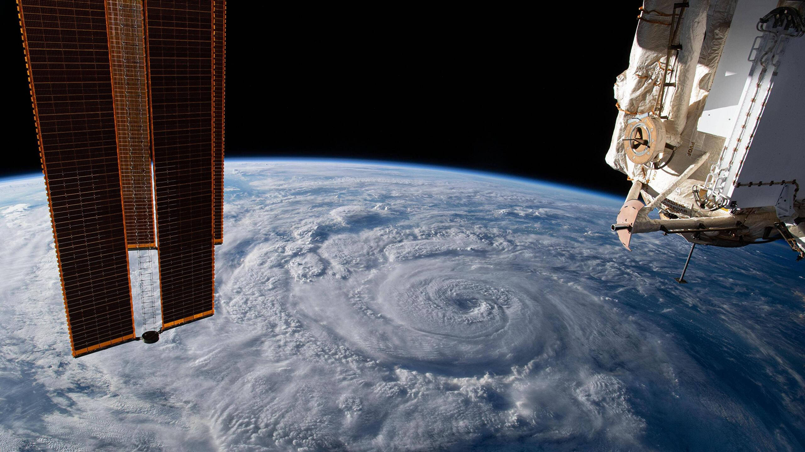 颶風衛星圖 壁紙 — 從國際太空站看到下方一個巨大颶風旋轉，前景有太陽能板和機械臂。