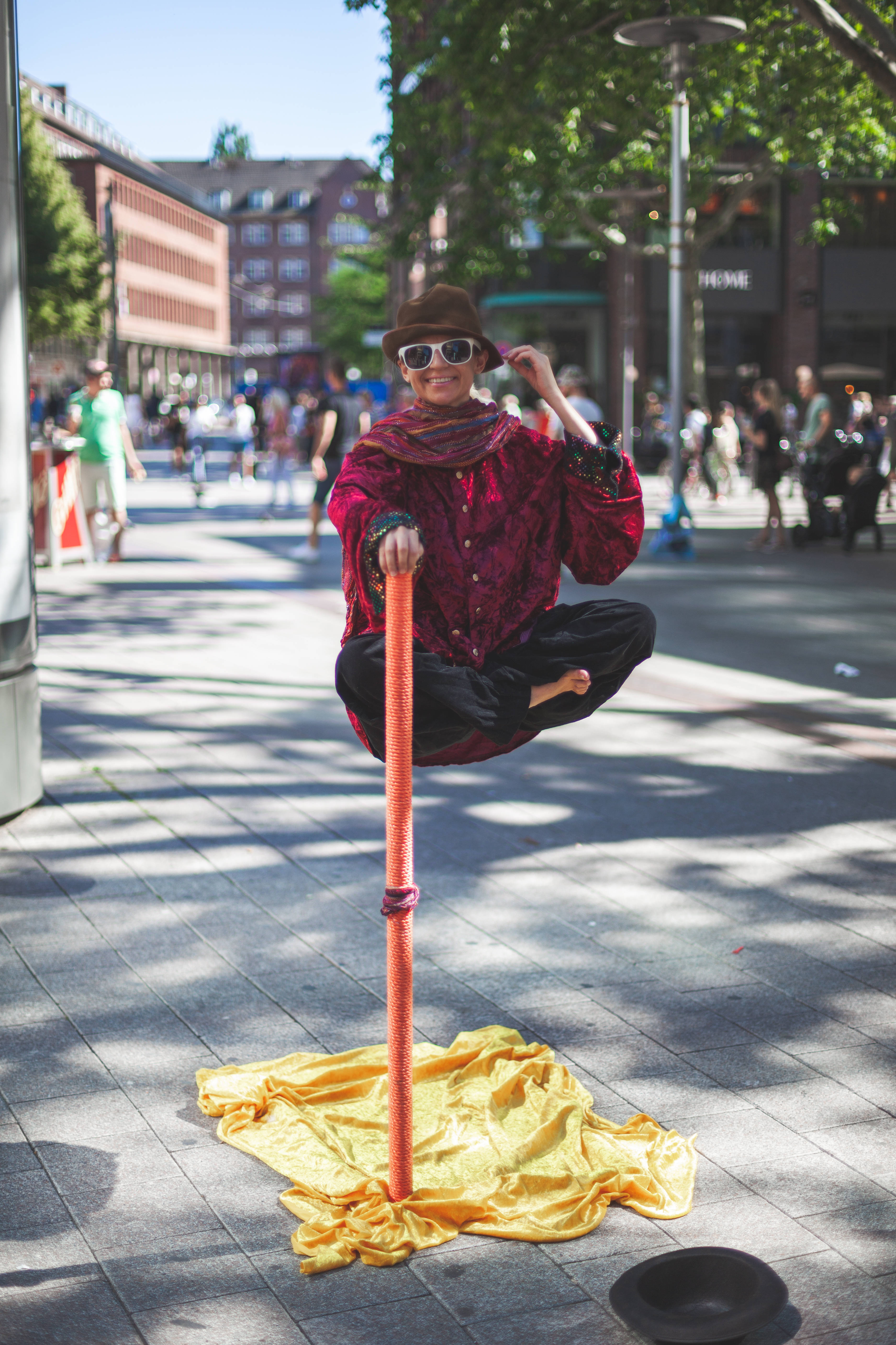 飄浮男子幻覺 壁紙 — 一位身穿紅色服裝和帽子的街頭表演者在陽光明媚的城市廣場上優雅地平衡在桿子上，同時對著鏡頭微笑。