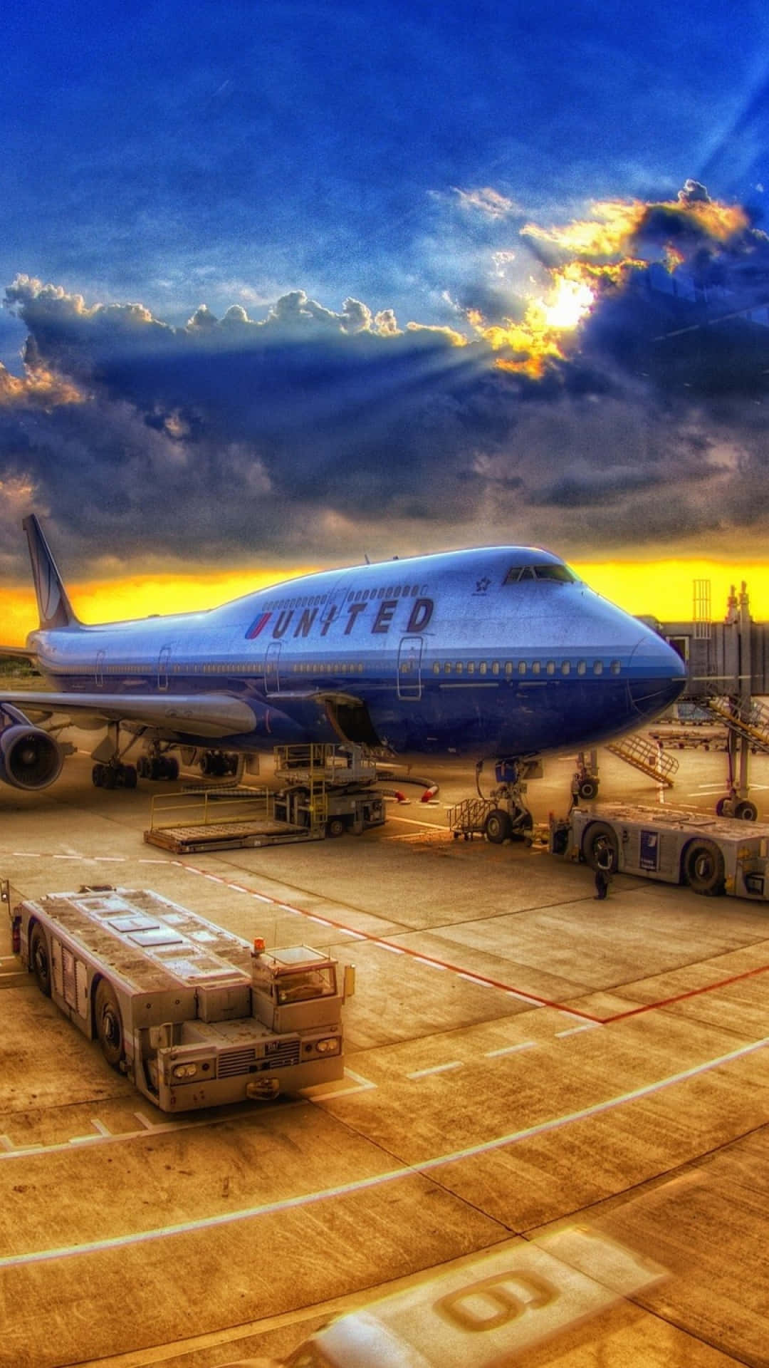 飛上雲霄，翱翔世界 壁紙 — 一架聯合航空珍寶客機停在機場閘口，背景是戲劇性的日落天空，陽光穿透雲層射出耀眼的光芒。