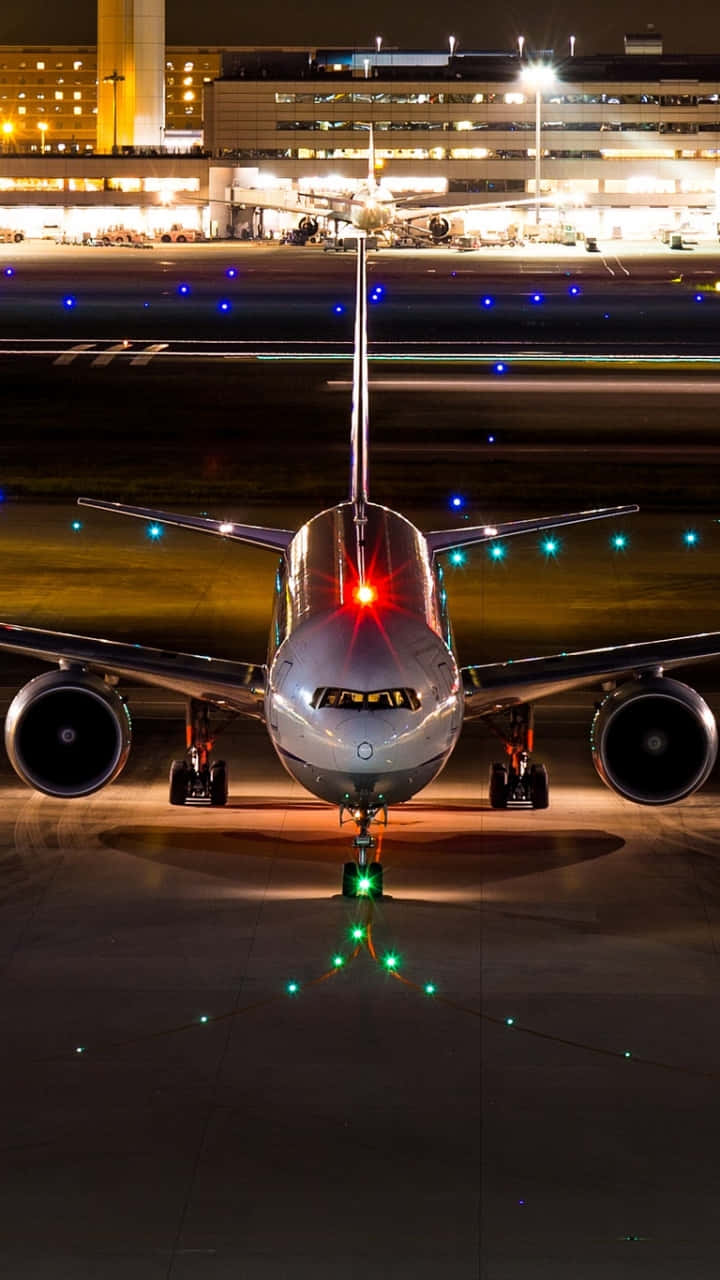 飛機停泊於跑道安卓飛機壁紙 壁紙 — 一架流線型飛機在夜間停在跑道上，背景有發光的燈光和明亮的客運大樓。
