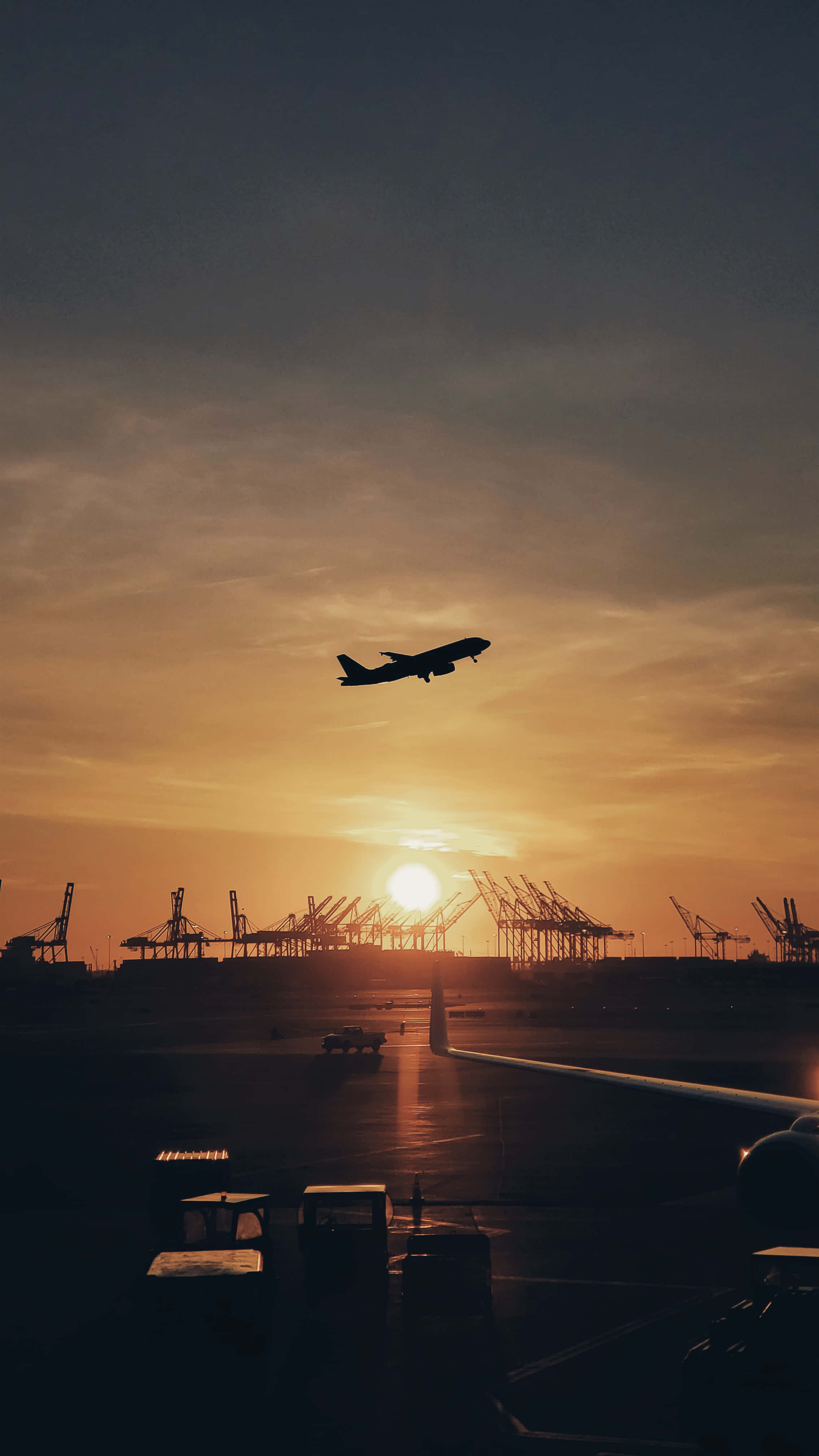 飛機剪影日落起飛離港 壁紙 — 一架飛機在金色夕陽的映襯下飛過機場，遠處有起重機。