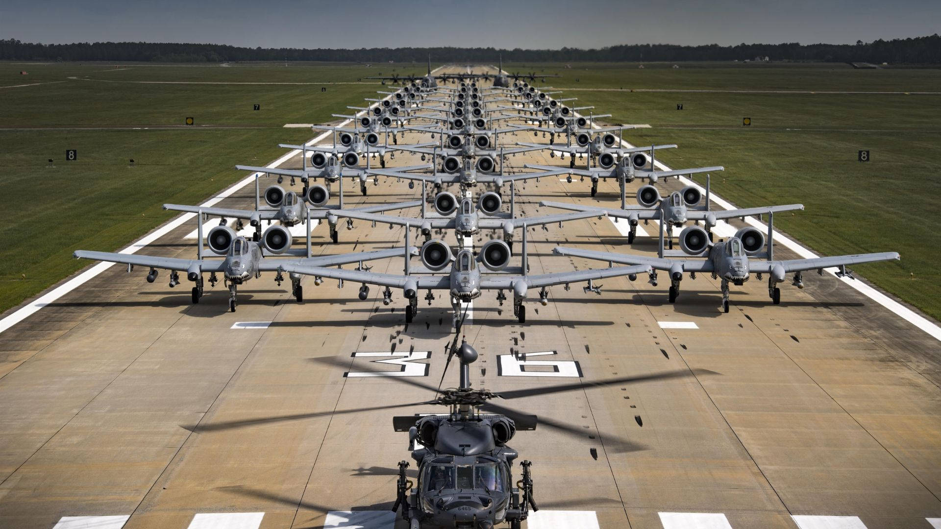 飛機填滿跑道 壁紙 — 從空中俯瞰眾多軍用攻擊機精確排列在停機坪上，前景有一架直升機。