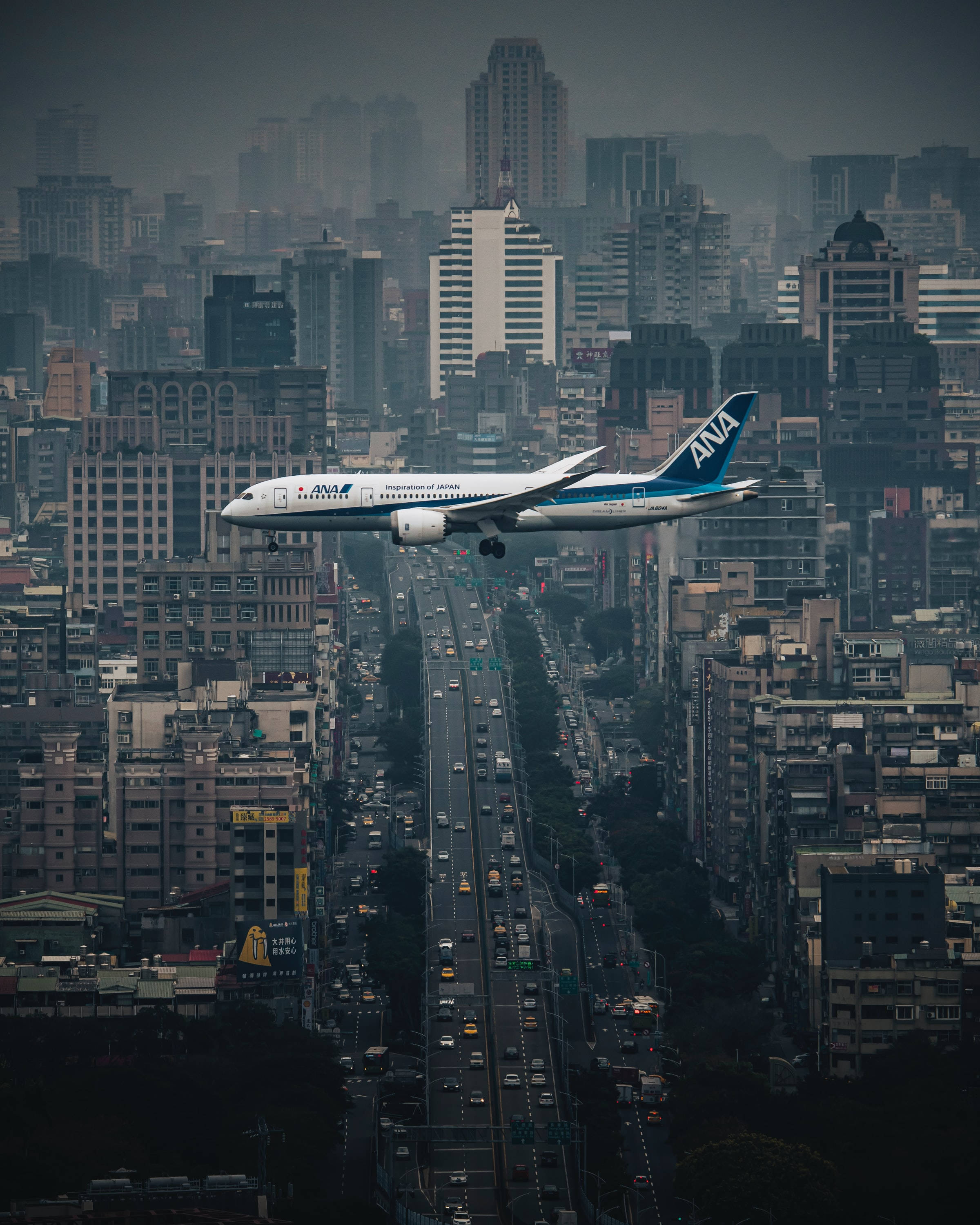 飛機掠過台北市 壁紙 — 一架全日空飛機在灰濛濛的天空下，低飛越過密集的城市天際線，下方是繁忙的高速公路。