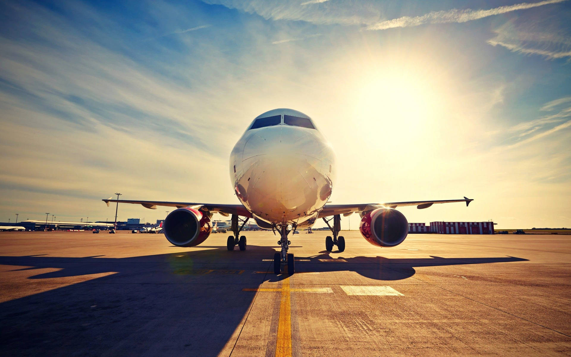 飛機於機場坡道與明亮陽光 壁紙 — 一架商用客機停在陽光明媚的停機坪上，太陽在其機頭後方閃耀，在跑道上投下長長的陰影。