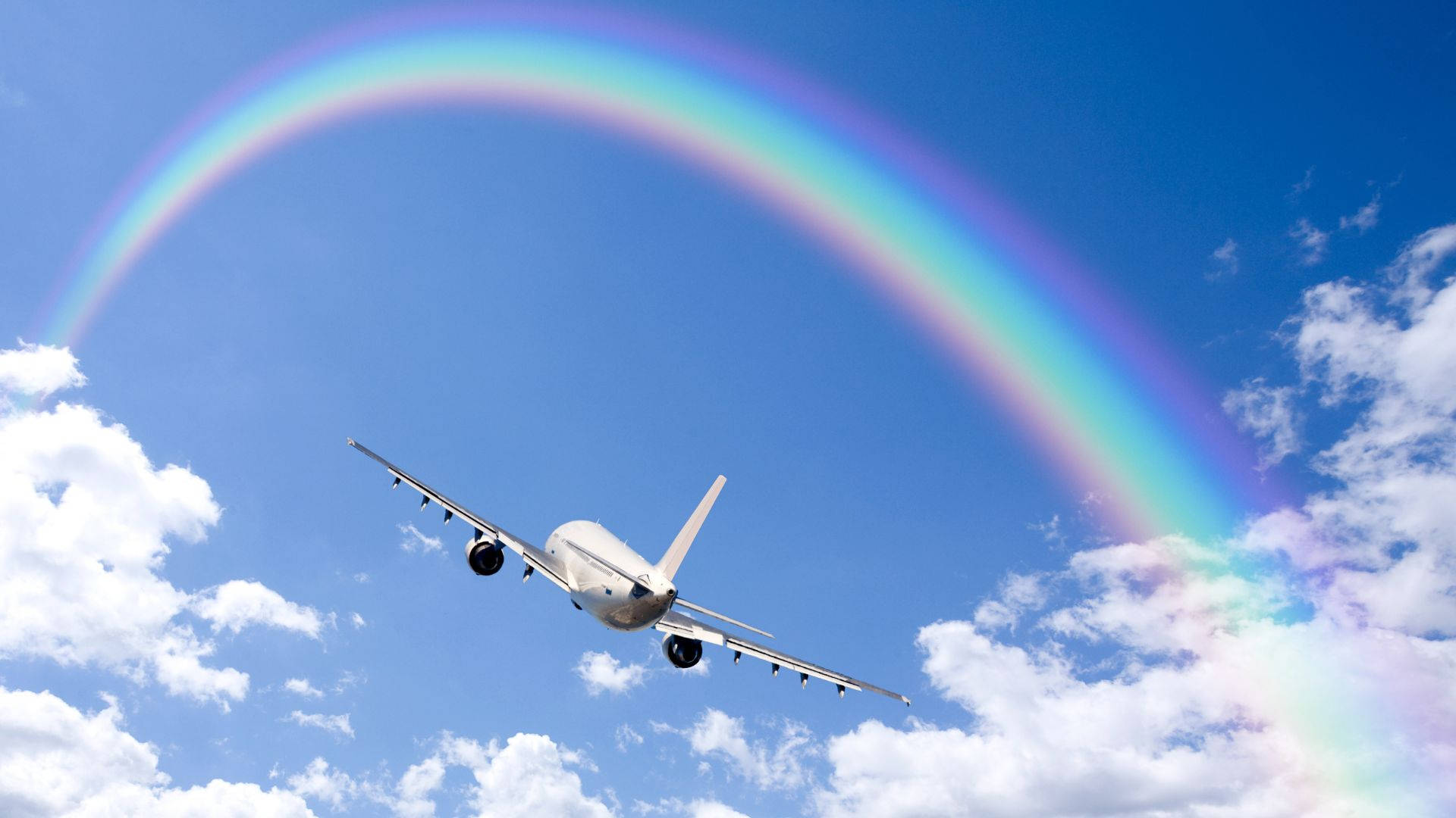 飛機穿越彩虹天空 壁紙 — 一架白色客機在橫跨明亮藍天、點綴著蓬鬆白雲的鮮豔彩虹下翱翔。