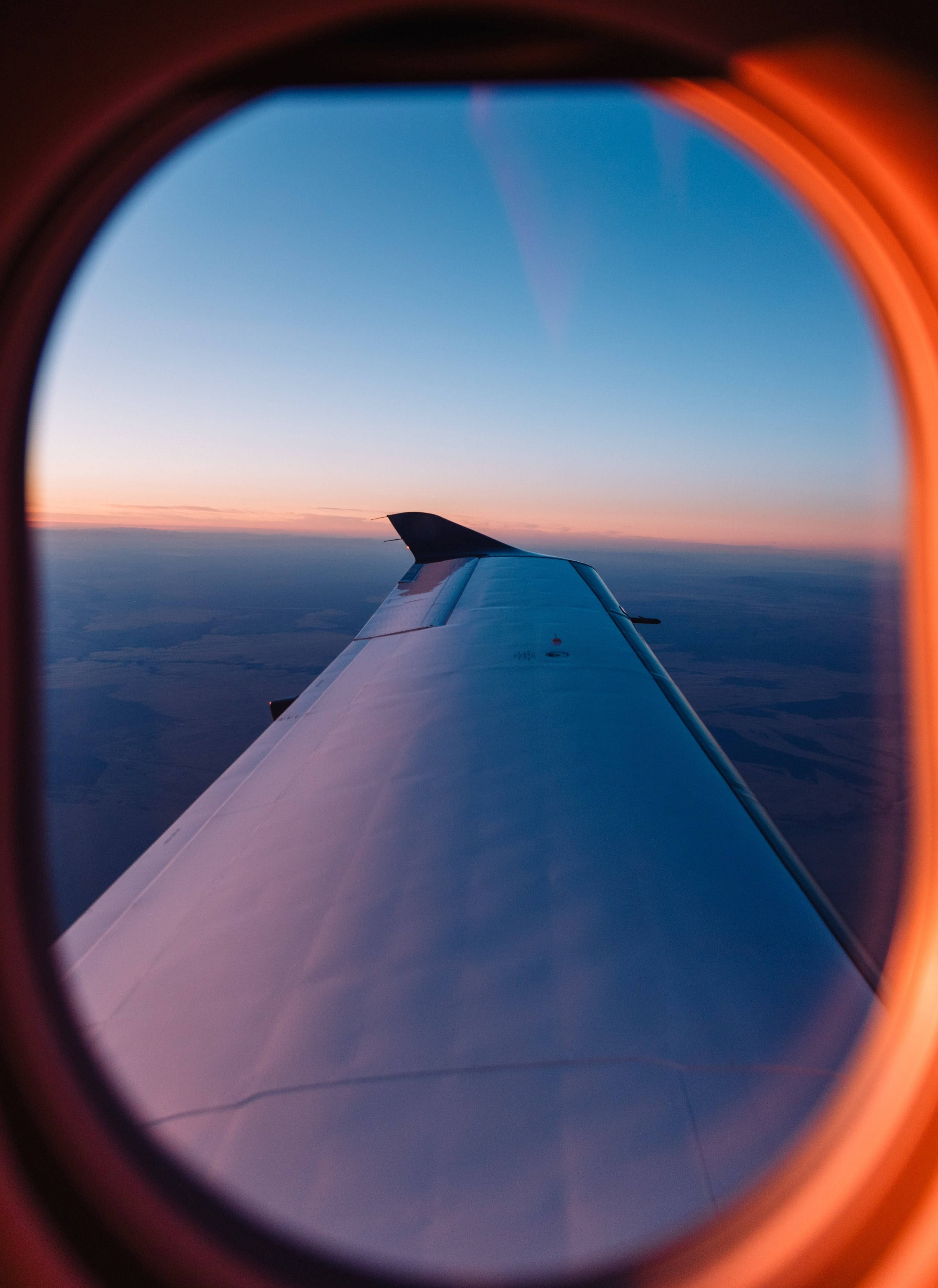飛機窗戶旅行唯美日落地平線 壁紙 — 透過圓形窗戶觀看的飛機機翼，在充滿活力的橙色和藍色天空的日落時分。