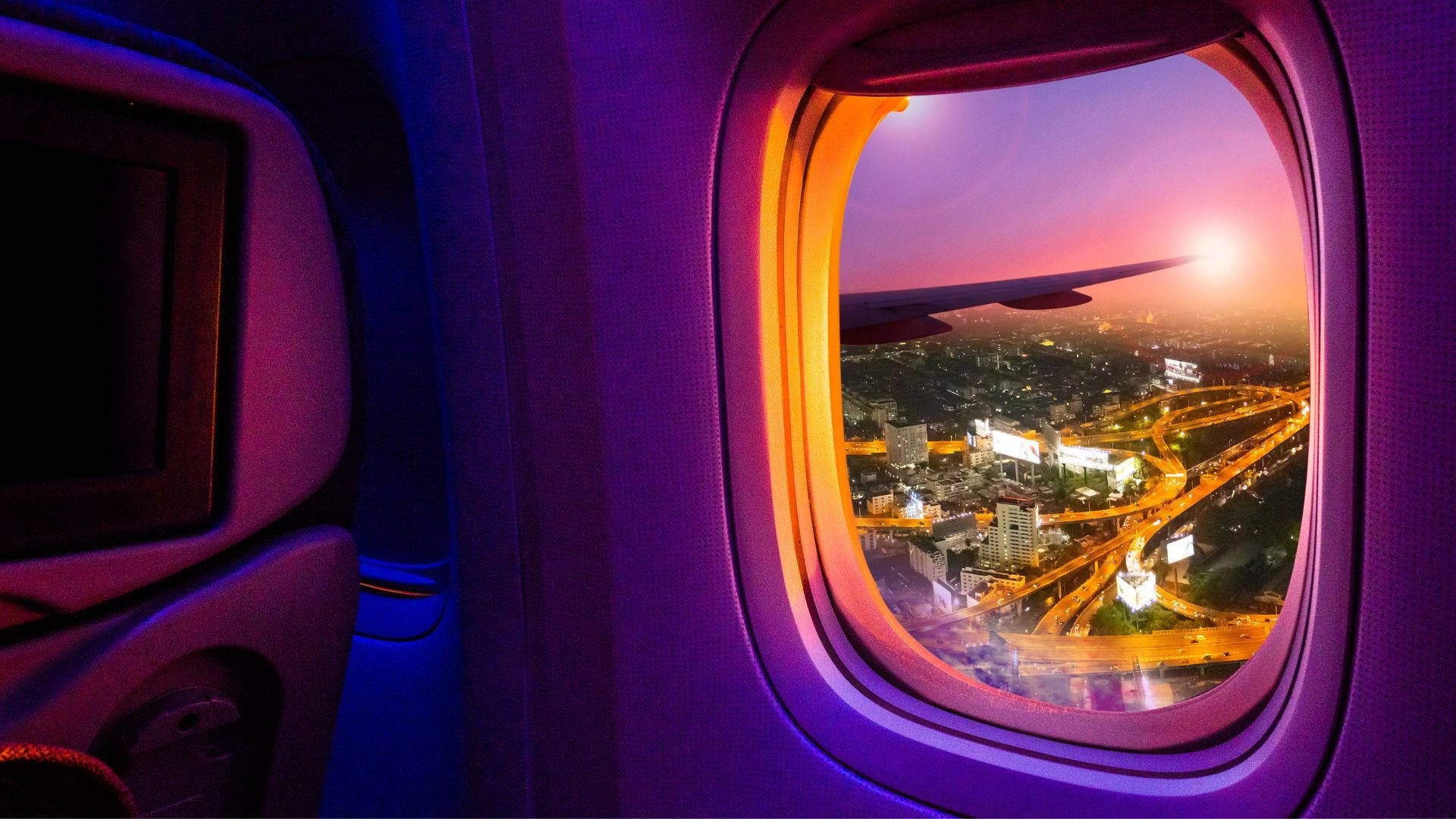 飛機窗戶旅行城市夜景 壁紙 — 日落時從飛機窗戶看到的景色，下方是發光的城市燈光和高速公路，帶有紫色內部照明。