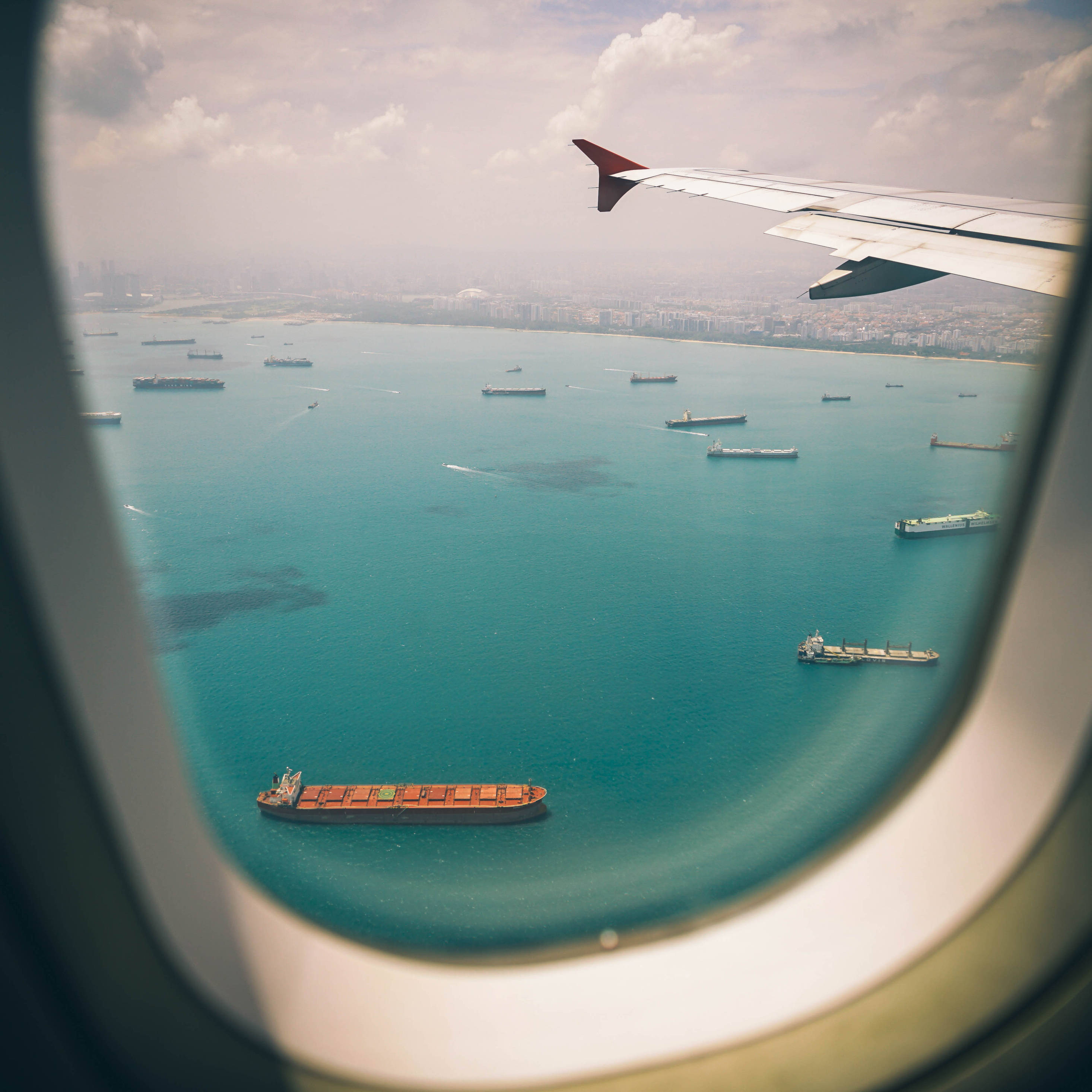 飛機窗戶旅行海景 壁紙 — 從飛機窗戶俯瞰，碧綠色的海面上點綴著貨船，遠處的城市景觀在陰天之下。