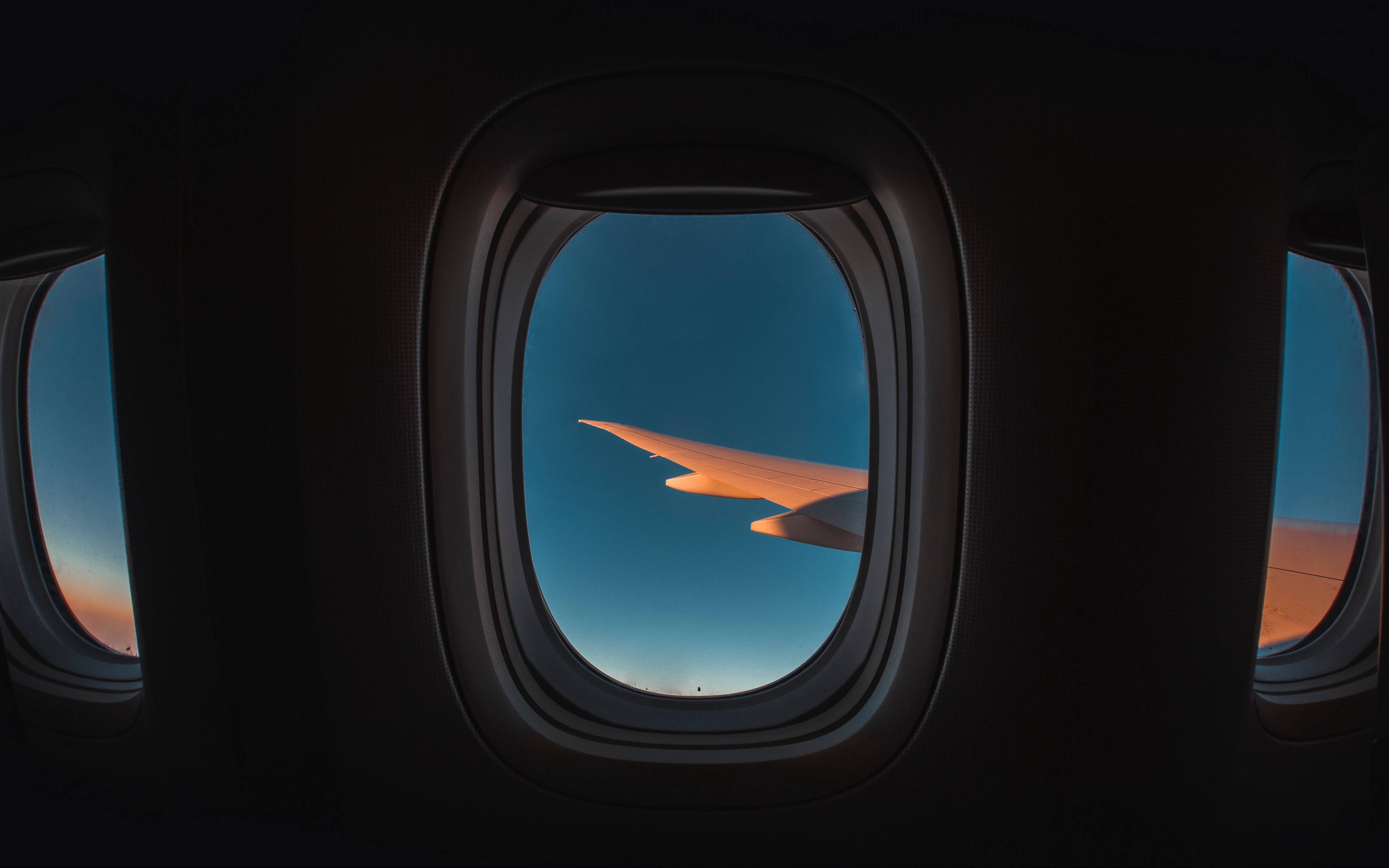 飛機窗戶旅行青綠色系天空 壁紙 — 飛機機翼在暮色天空中發出琥珀色光芒，透過飛機窗戶看到黑暗的機艙內部。