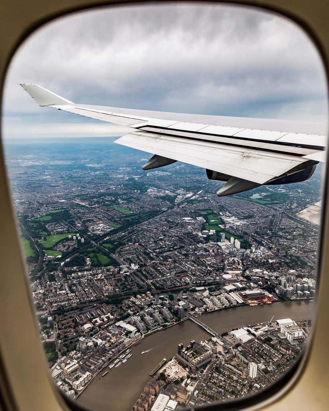 飛機窗景 壁紙 — 透過飛機窗戶俯瞰，機翼下方是廣闊的城市景觀，河流和橋樑在陰天之下。