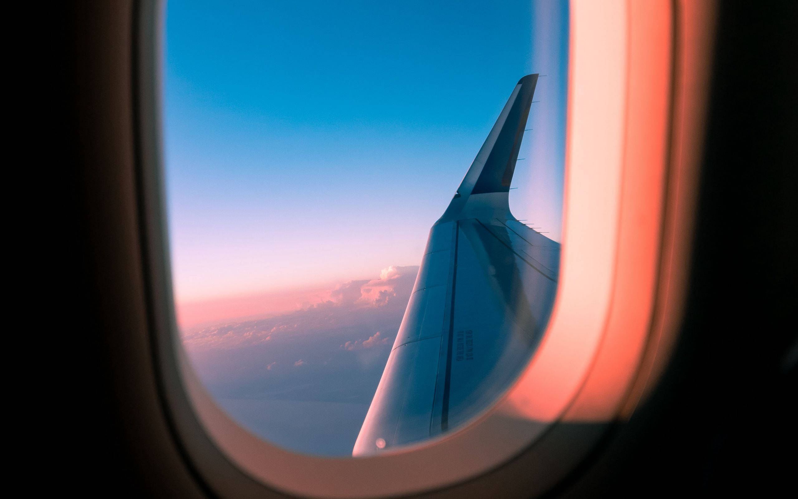飛機窗邊令人屏息的天空景色 壁紙 — 透過飛機窗戶看到的飛機機翼，日落時分，粉紅色和藍色的天空，下方有雲。