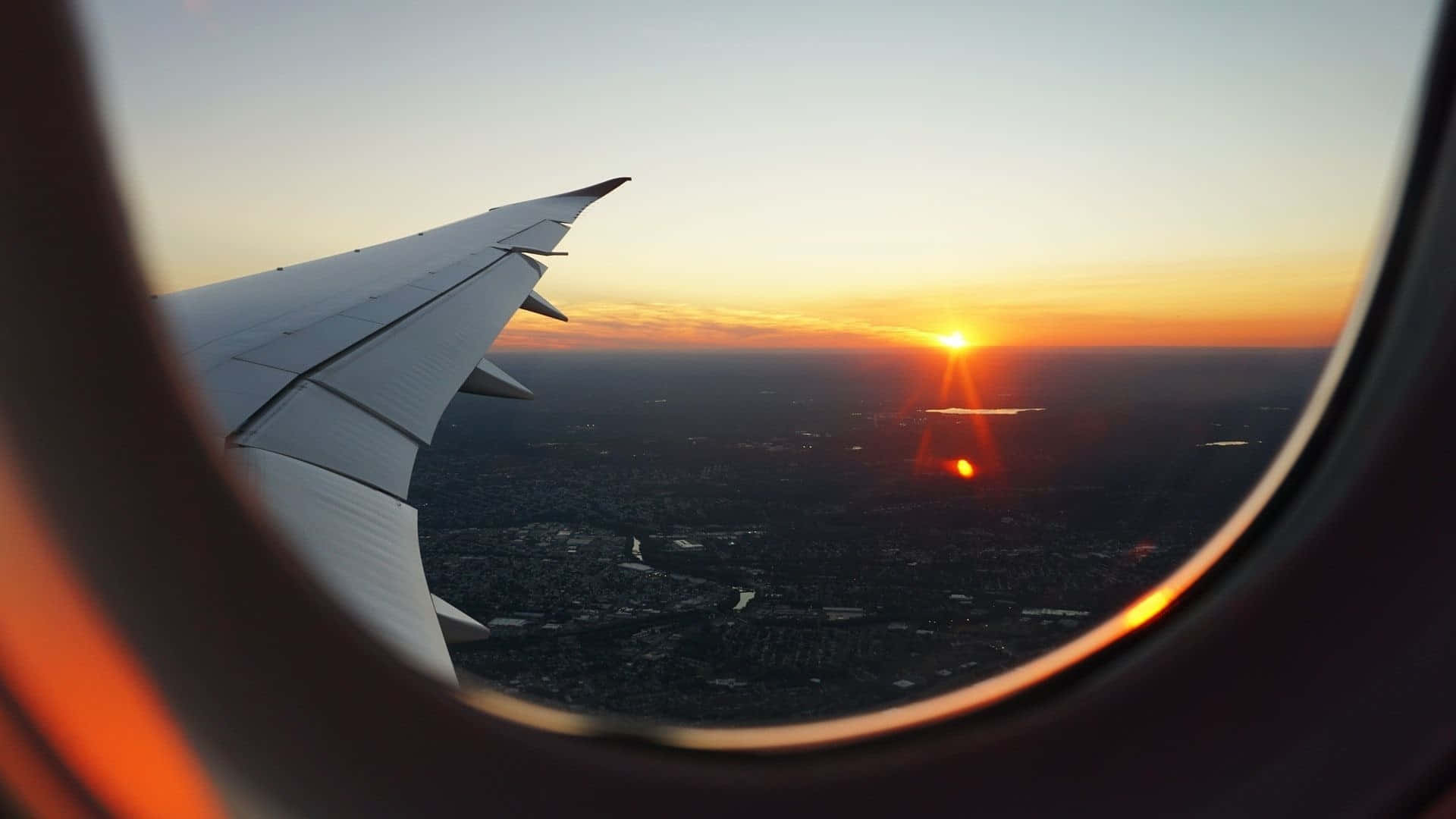飛機窗邊日落與機翼離港 壁紙 — 從飛機窗戶看到的日落，機翼剪影映襯著橙色天空，下方是廣闊的城市景觀。