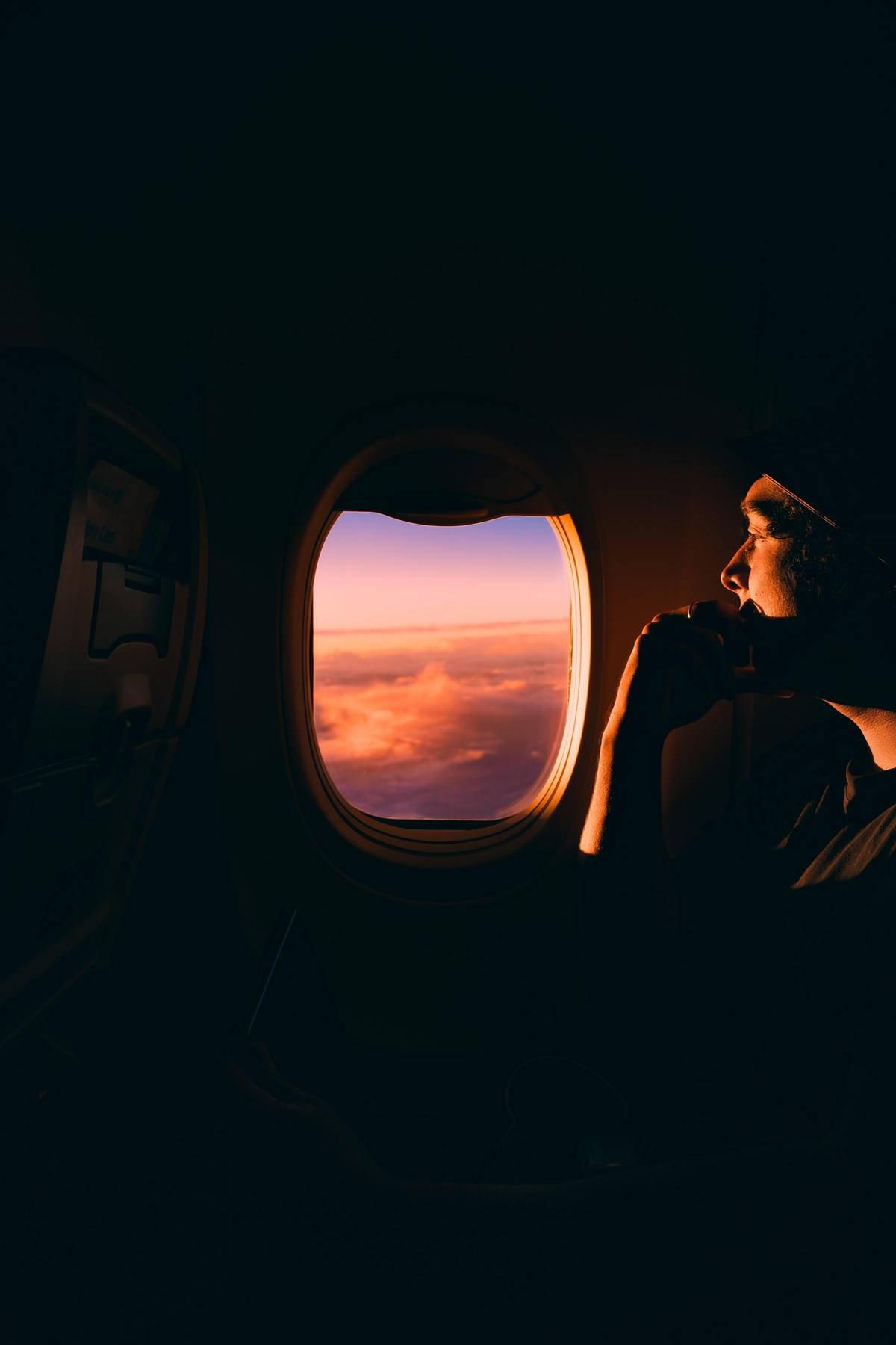 飛機窗邊的乘客欣賞夕陽天空 壁紙 — 一個人從飛機窗戶凝視著蓬鬆雲層上方充滿活力的橙色和粉紅色日落。