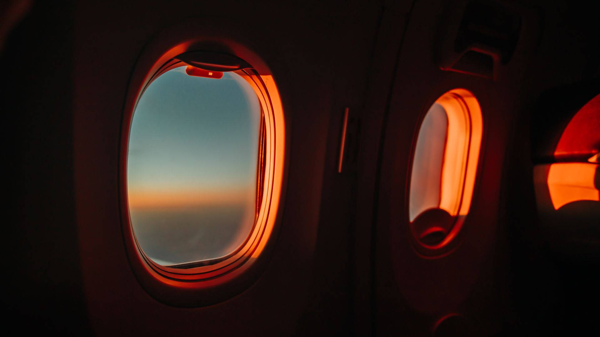 飛機窗邊的唯美日落天空 壁紙 — 飛機客艙窗戶框住寧靜的日落，橙色光芒映襯著柔和的藍天。