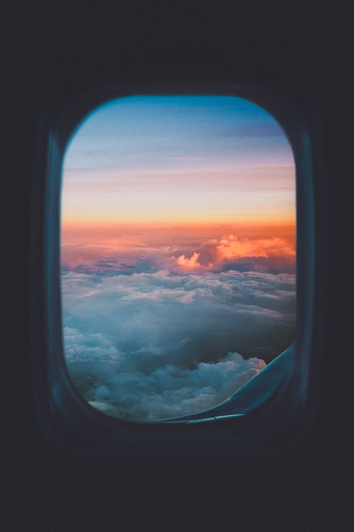 飛機窗邊的夕陽雲景 壁紙 — 飛機窗戶框住雲層上令人驚嘆的日落，呈現橙、粉紅和藍色調。
