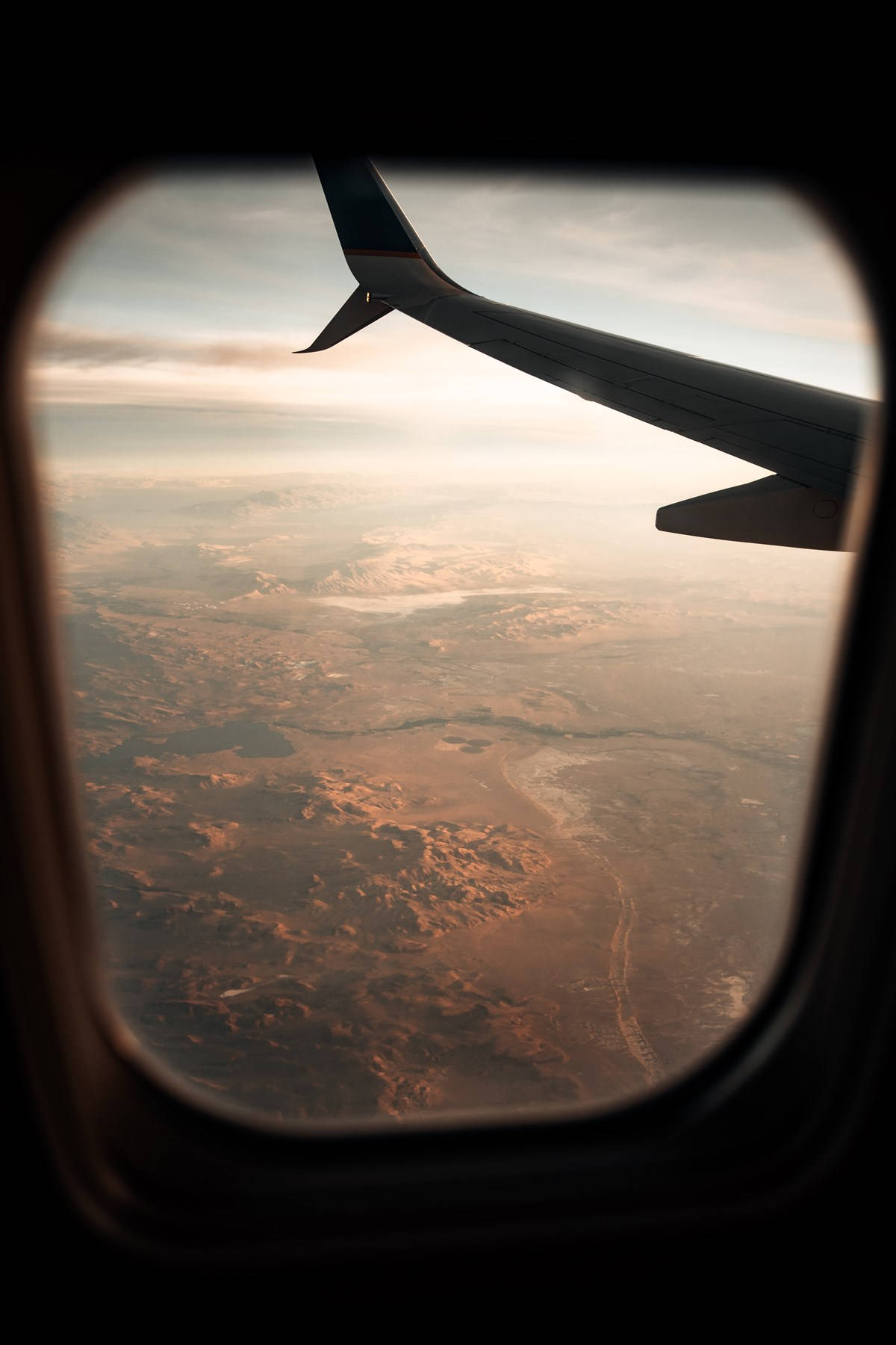飛機窗邊的復古夕陽風景 壁紙 — 從飛機窗戶看到的飛機翼剪影，框住了崎嶇沙漠地形上的金色日落。