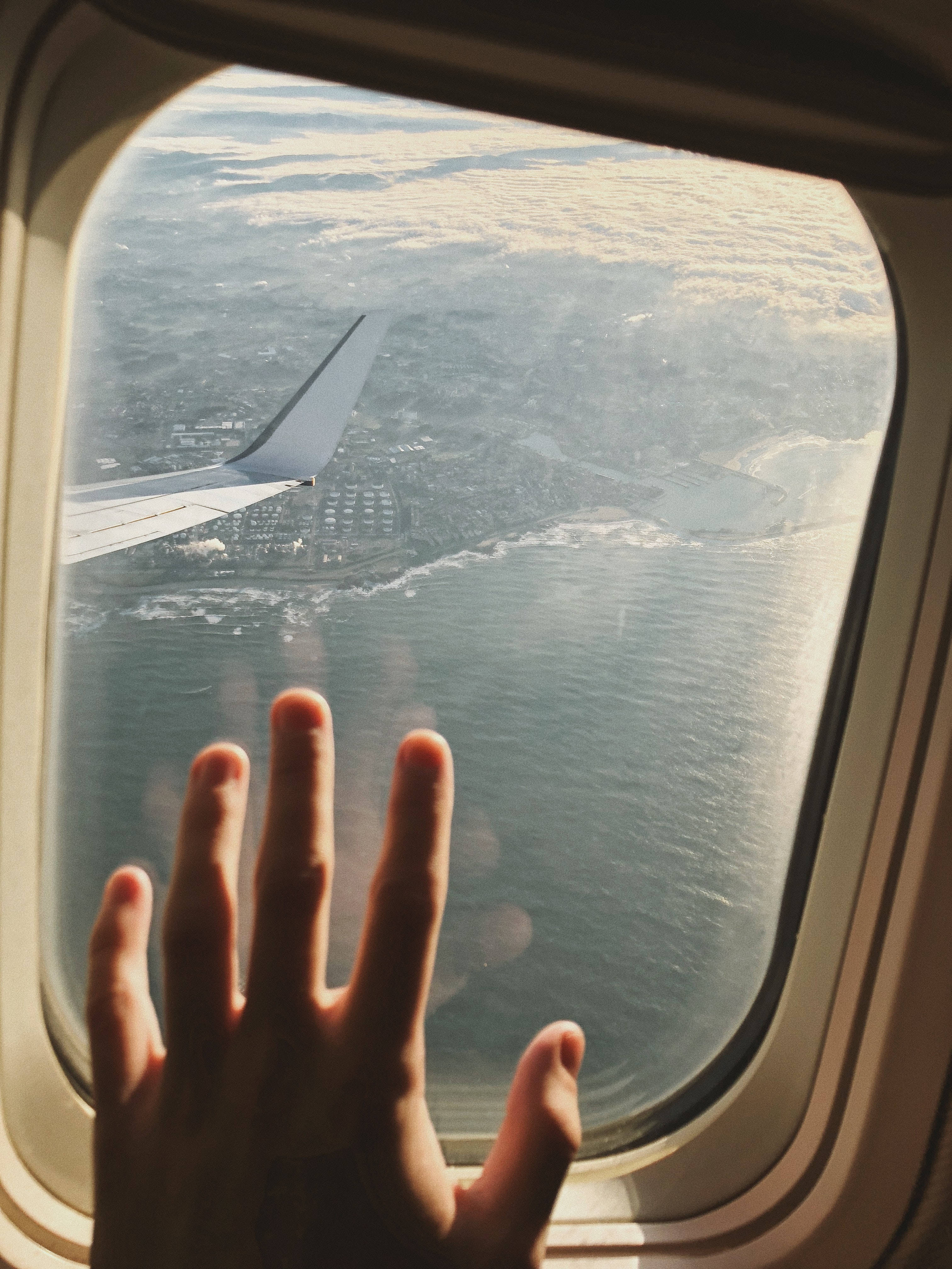 飛機窗邊的手與旅行渴望 壁紙 — 一隻手按在飛機窗上，機翼延伸至沿海城市和下方的海洋。