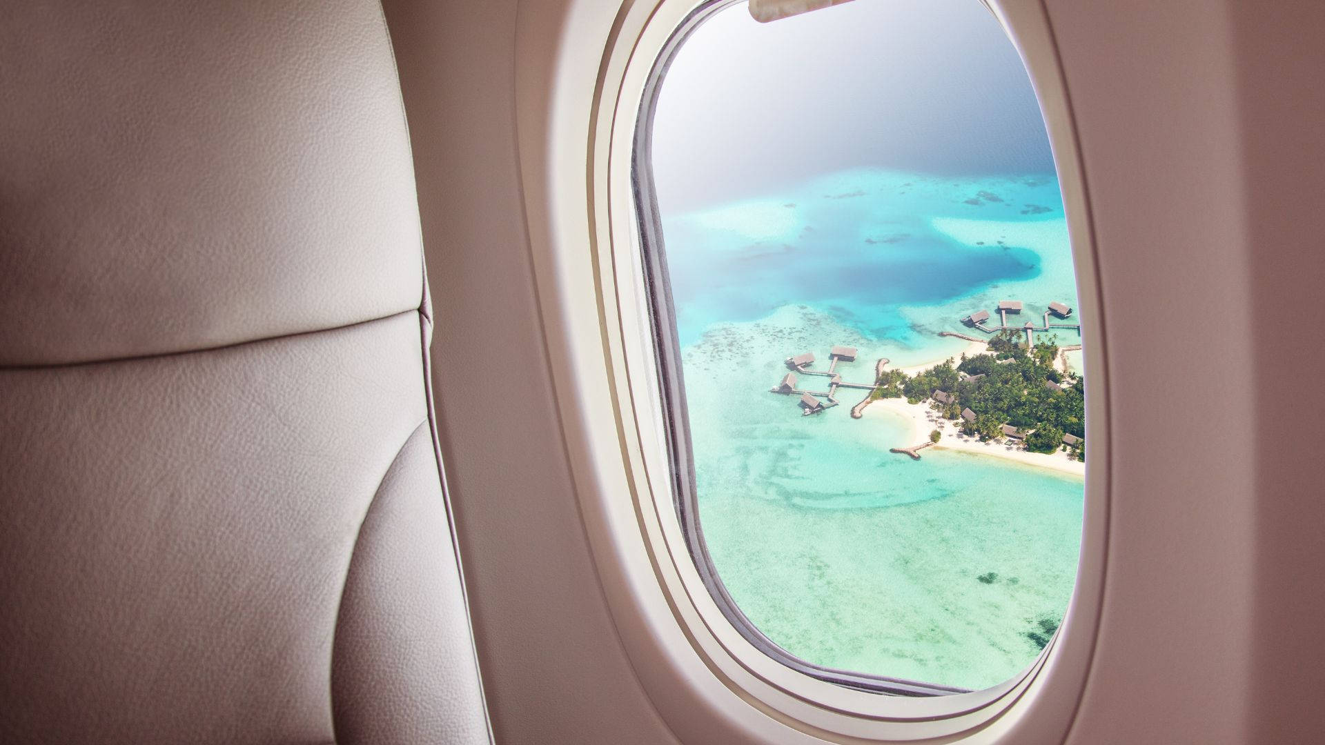 飛機窗邊的涼爽島嶼風景 壁紙 — 從飛機窗戶俯瞰，呈現碧綠潟湖、水上平房和下方白色沙灘的景色。