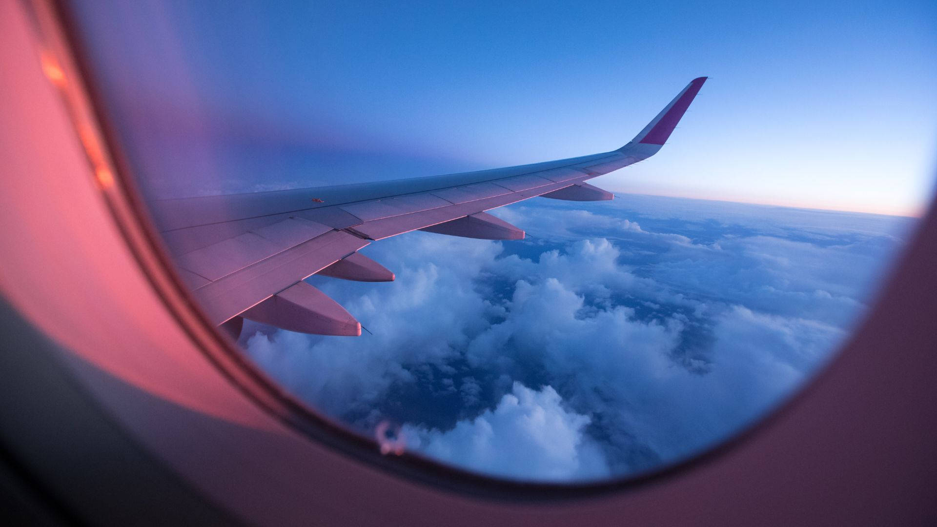 飛機窗邊的藍色雲海風景 壁紙 — 透過飛機窗戶在黃昏時分看到的飛機機翼，粉紅色和藍色的天空在蓬鬆的雲層之上。