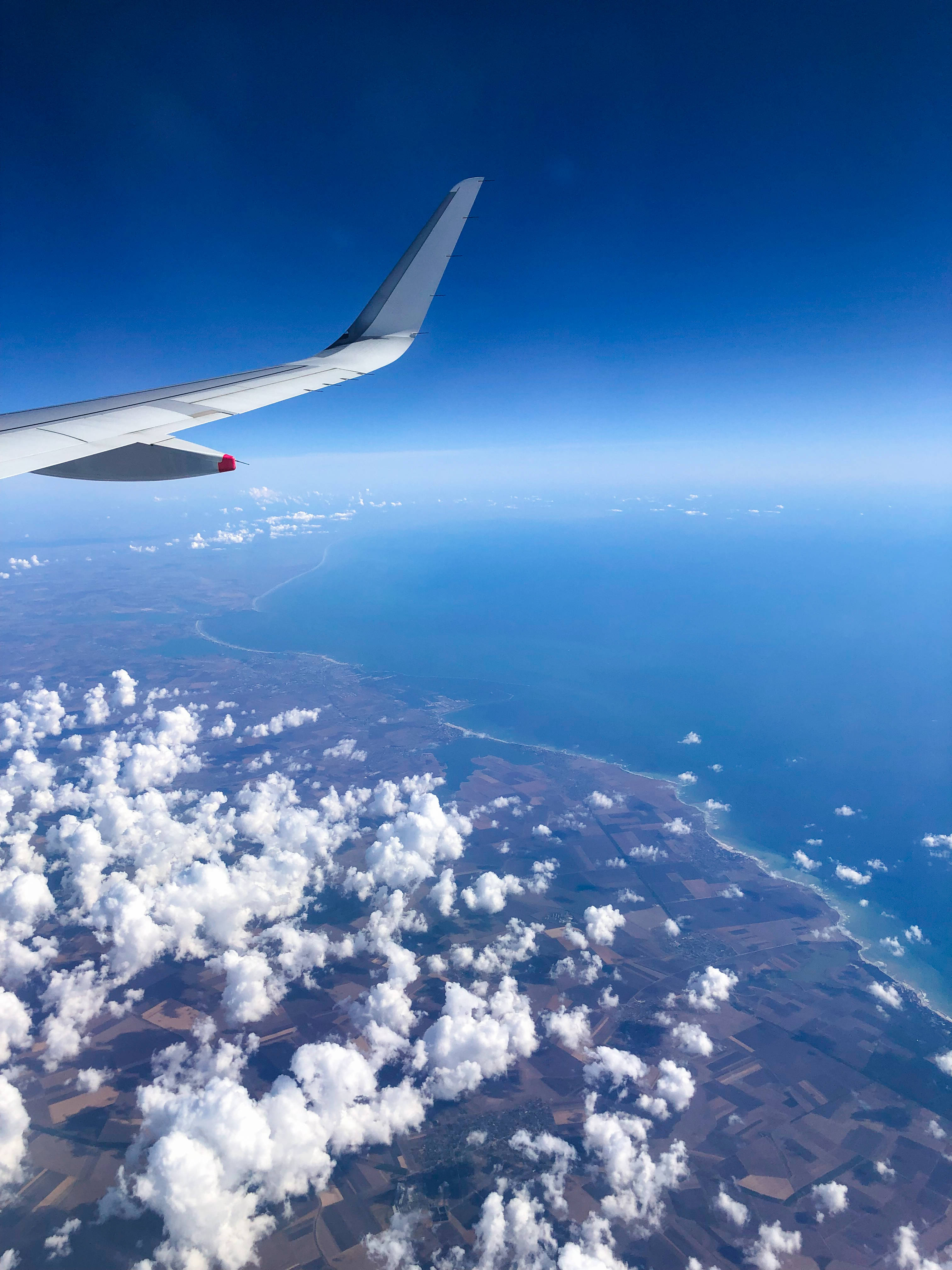 飛機翅膀空中視角 iPhone 壁紙 — 從飛機窗戶俯瞰，機翼在蓬鬆雲層之上，下方是海岸景觀，天空清澈湛藍。