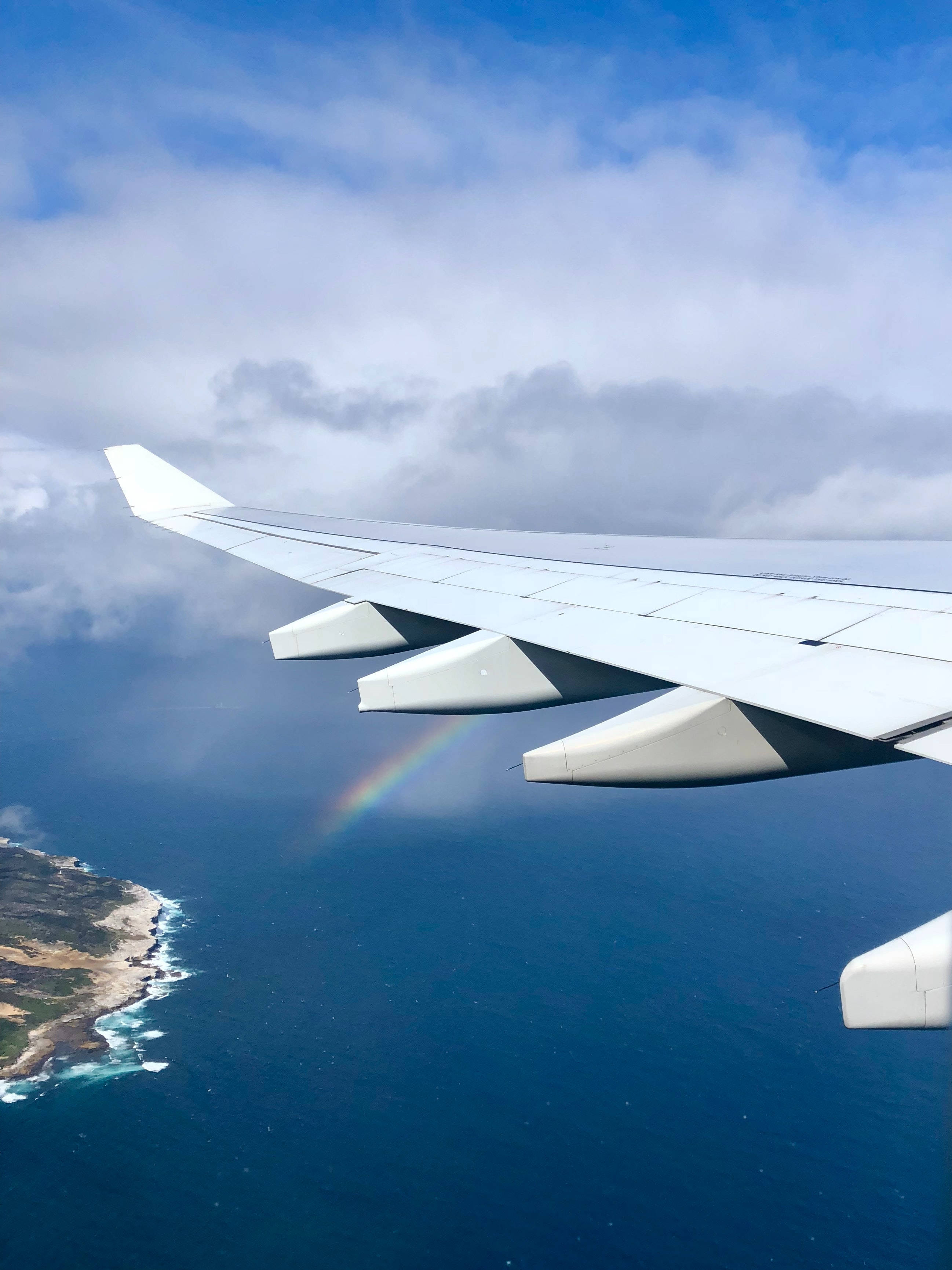 飛機翼下彩虹 壁紙 — 飛機機翼延伸至海洋上方，一道鮮豔的彩虹在明亮的藍天下穿過雲層彎曲。