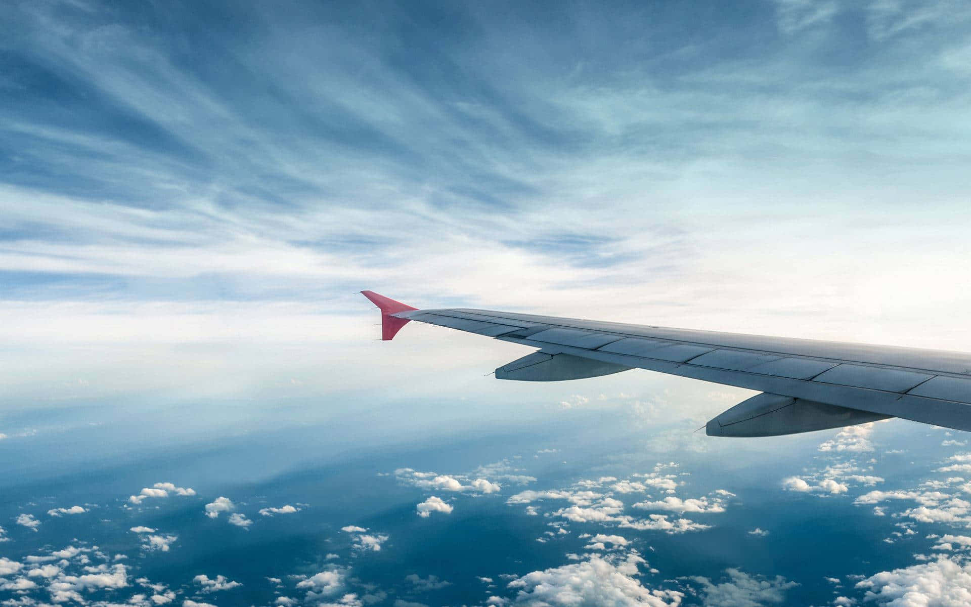 飛機翼在藍天雲層背景 壁紙 — 飛機機翼延伸在蓬鬆的白雲之上，下方是廣闊的藍天，點綴著縷縷卷雲。