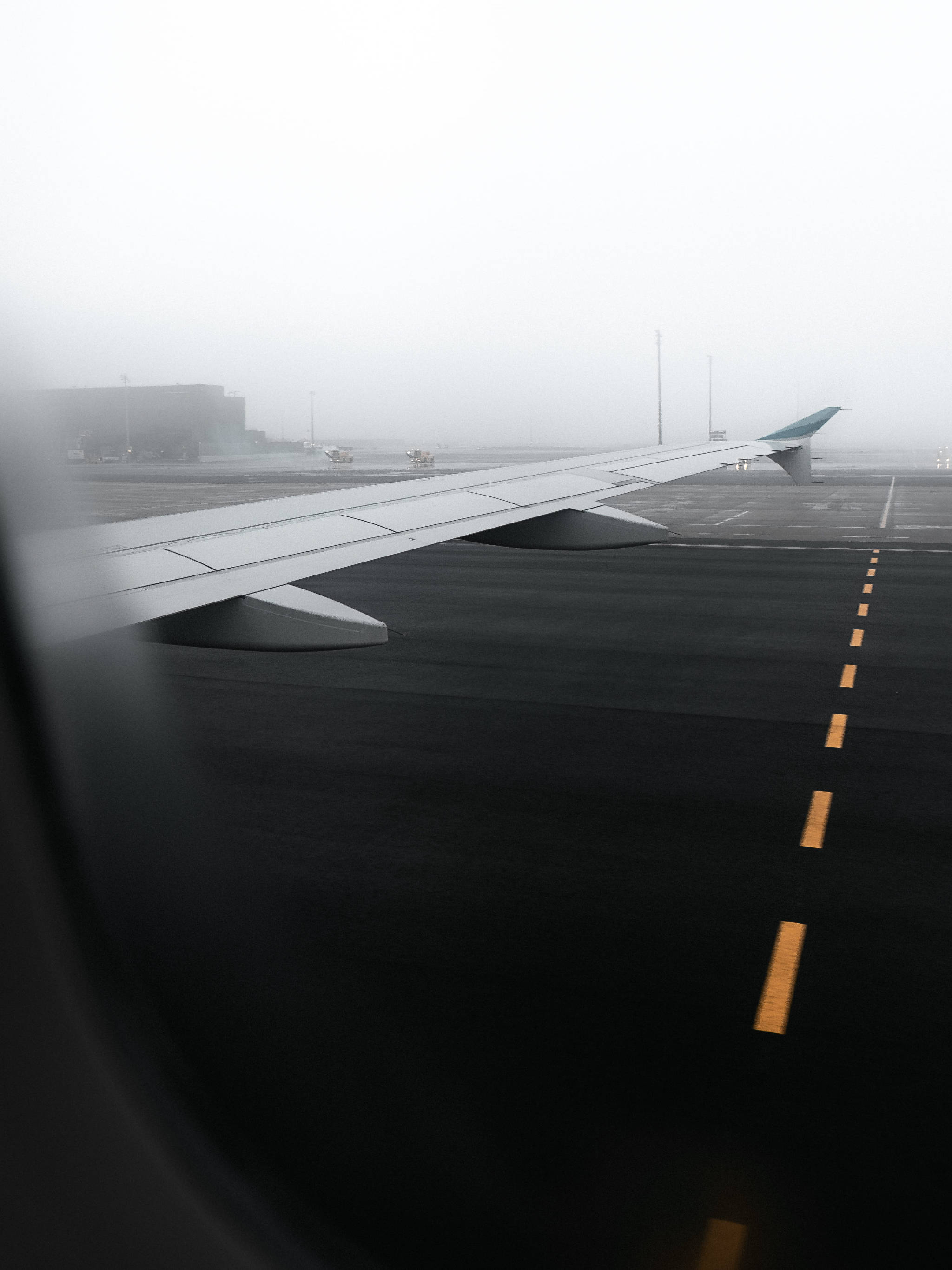 飛機翼掠過跑道 壁紙 — 從飛機窗戶看到的霧氣瀰漫的機場停機坪，機翼延伸在黃色跑道標記上方。