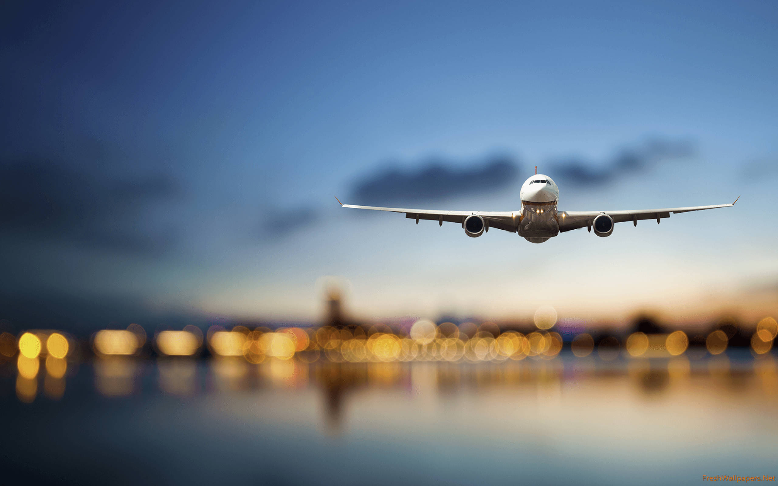 飛機與模糊城市燈光 壁紙 — 一架大型客機在黃昏時低飛過水面，下方模糊的城市燈光倒映著。