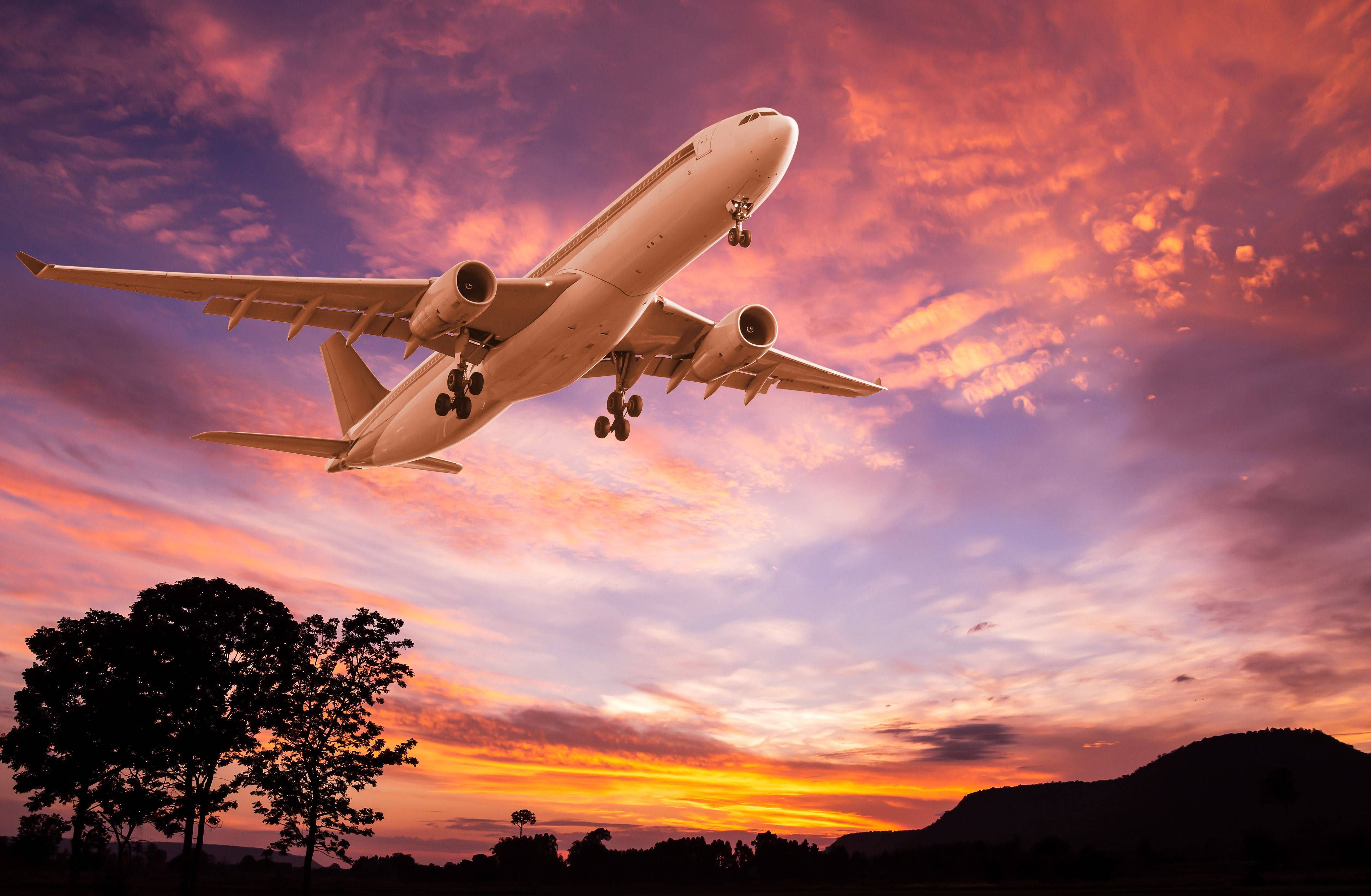 飛機與漸層粉紅天空 壁紙 — 一架大型客機在火紅的日落天空中攀升，下方是剪影般的樹木和山丘。