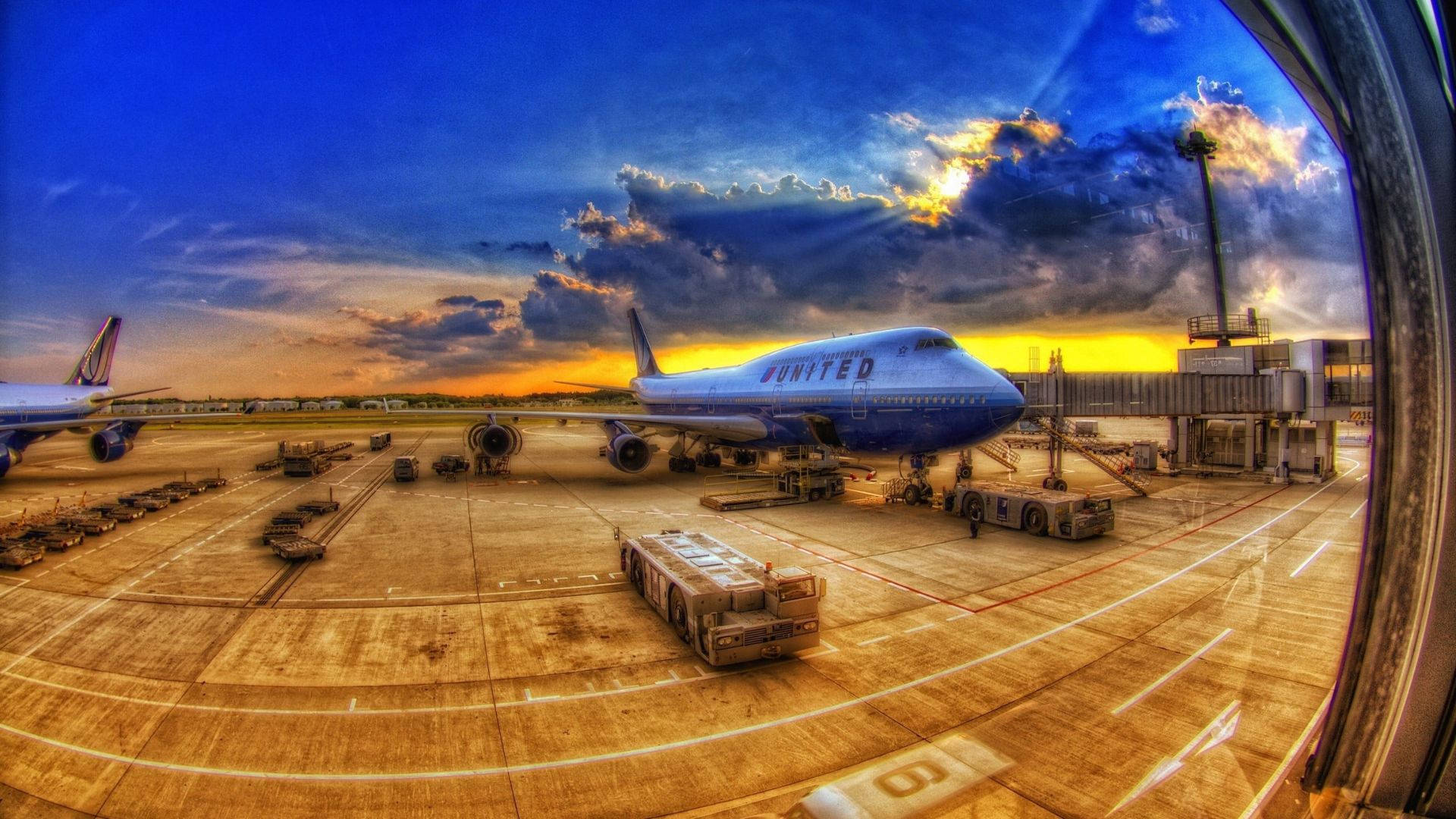 飛機與貨物停泊於跑道 壁紙 — 一架聯合航空珍寶客機在充滿活力的日落時分停靠在機場閘口，伴隨著戲劇性的雲層和魚眼鏡頭變形。