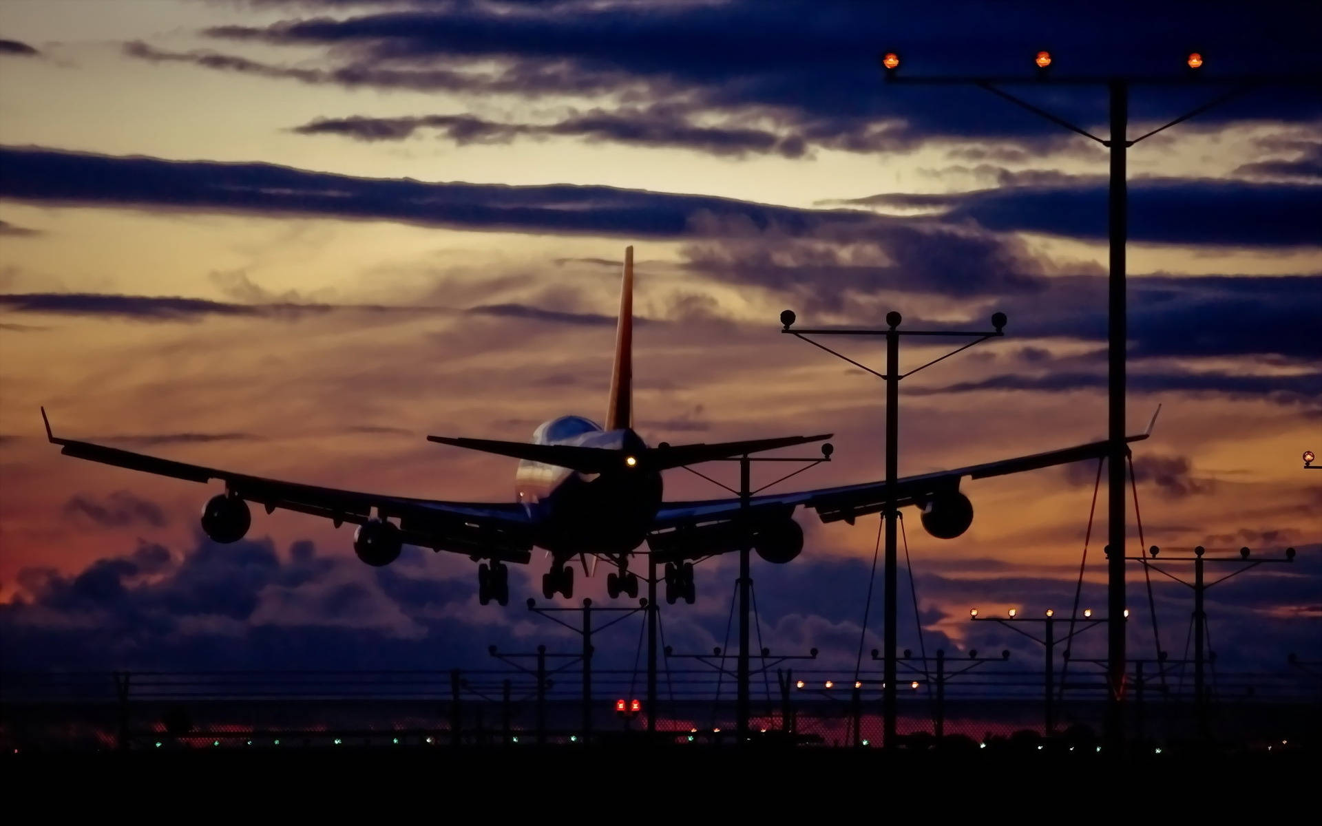 飛機降落機場 壁紙 — 一架大型飛機在戲劇性的黃昏天空襯托下呈現剪影，天空帶有橙色和紫色的雲彩條紋。