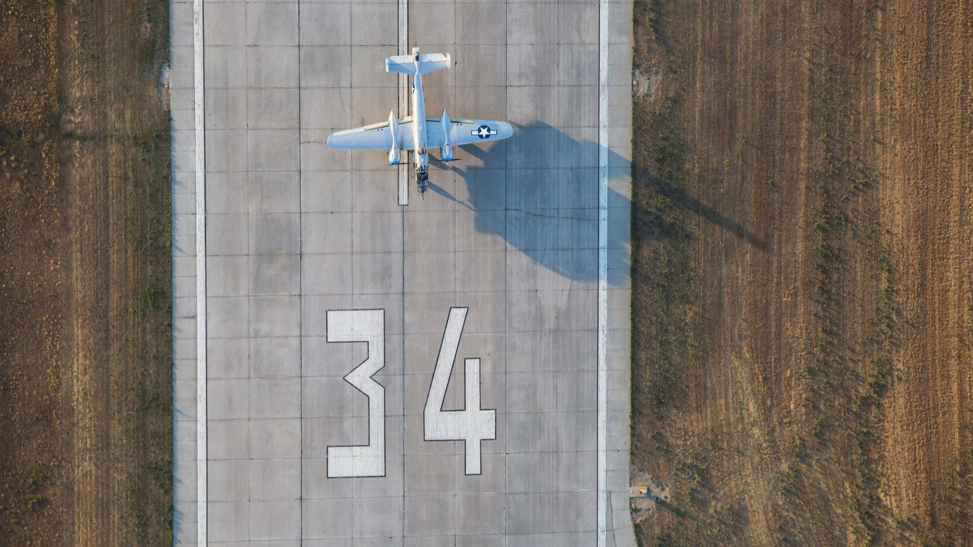 飛機降落跑道 壁紙 — 從空中俯瞰一架復古軍用飛機停在34號跑道上，在混凝土上投下長長的影子。