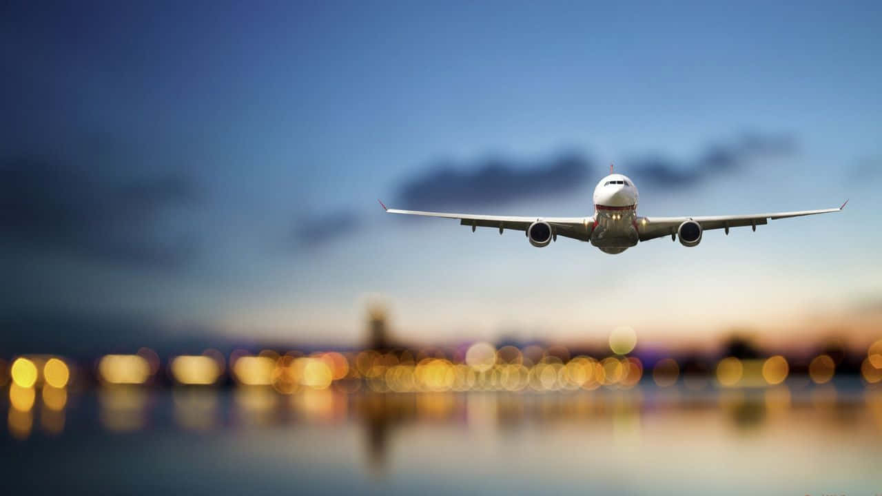 飛機飛越色彩繽紛天空的驚豔空中視角 壁紙 — 一架商用飛機在暮色天空中攀升，下方水面倒映著模糊的城市燈光。