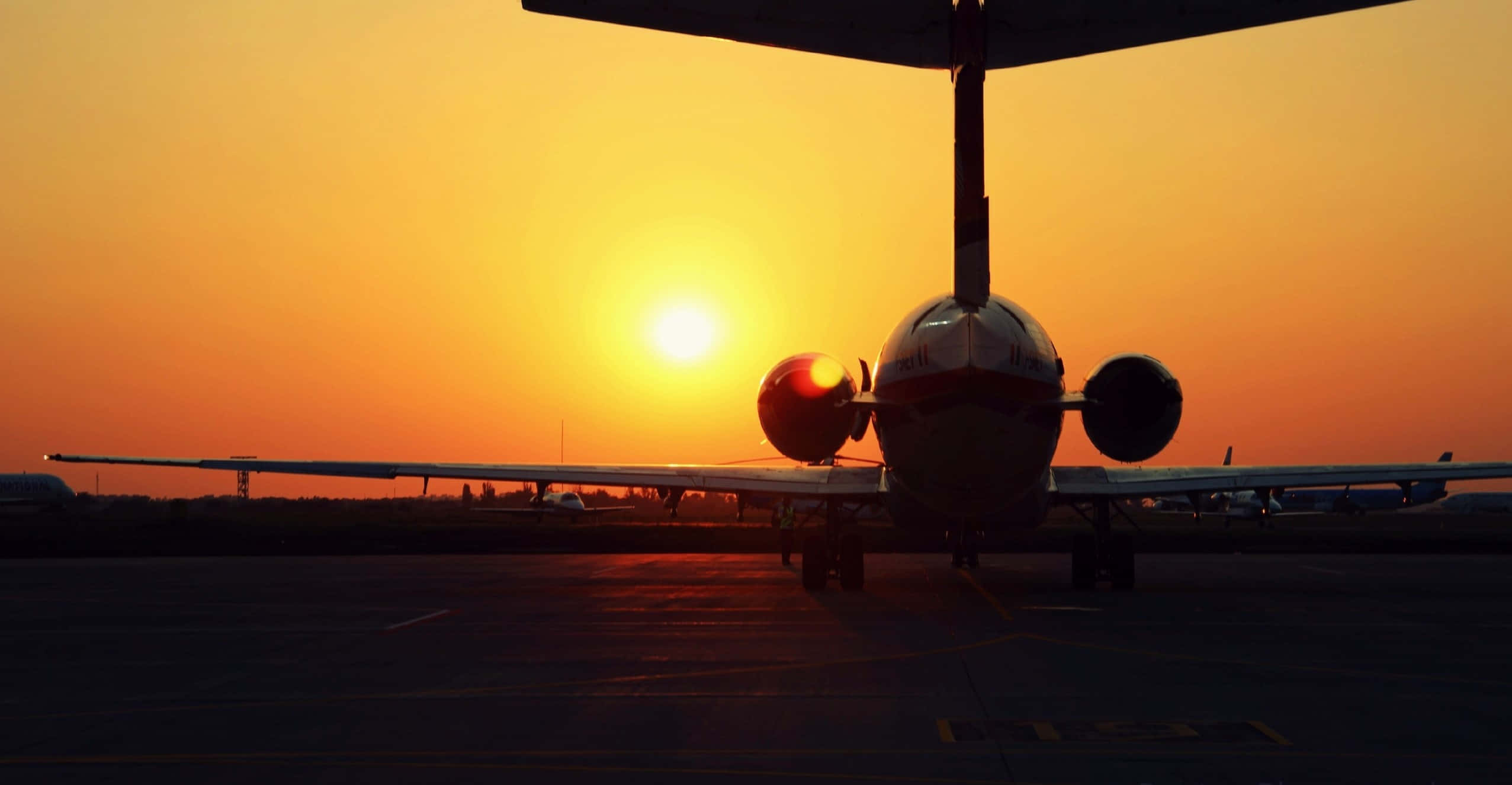 飛機飛越藍天 壁紙 — 一架雙引擎飛機在機場停機坪上，以火紅橙色日落為背景的剪影。