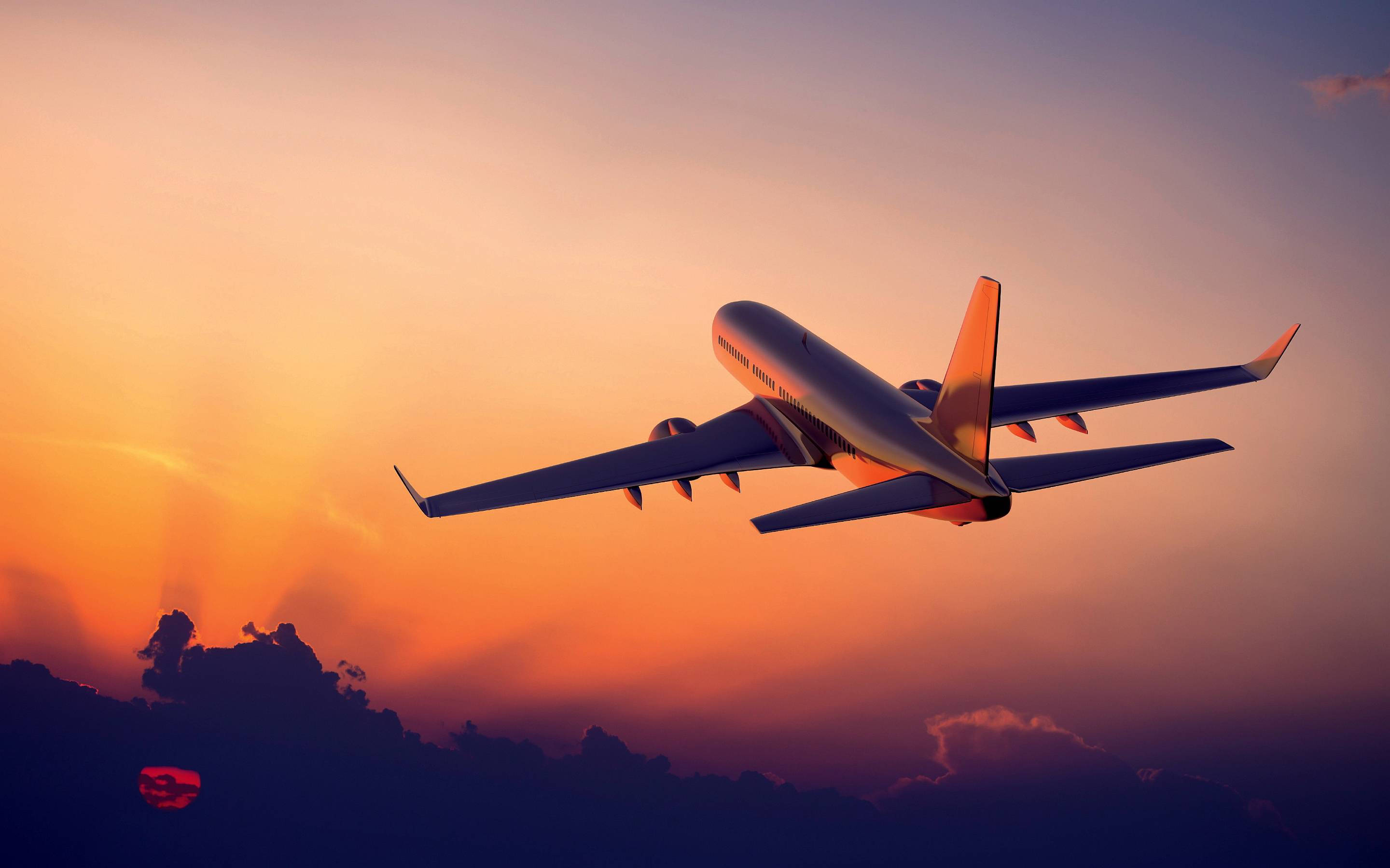 飛行中的飛機與夕陽 壁紙 — 一架商用飛機在充滿活力的橙紫色調日落天空和下方剪影雲朵中翱翔。
