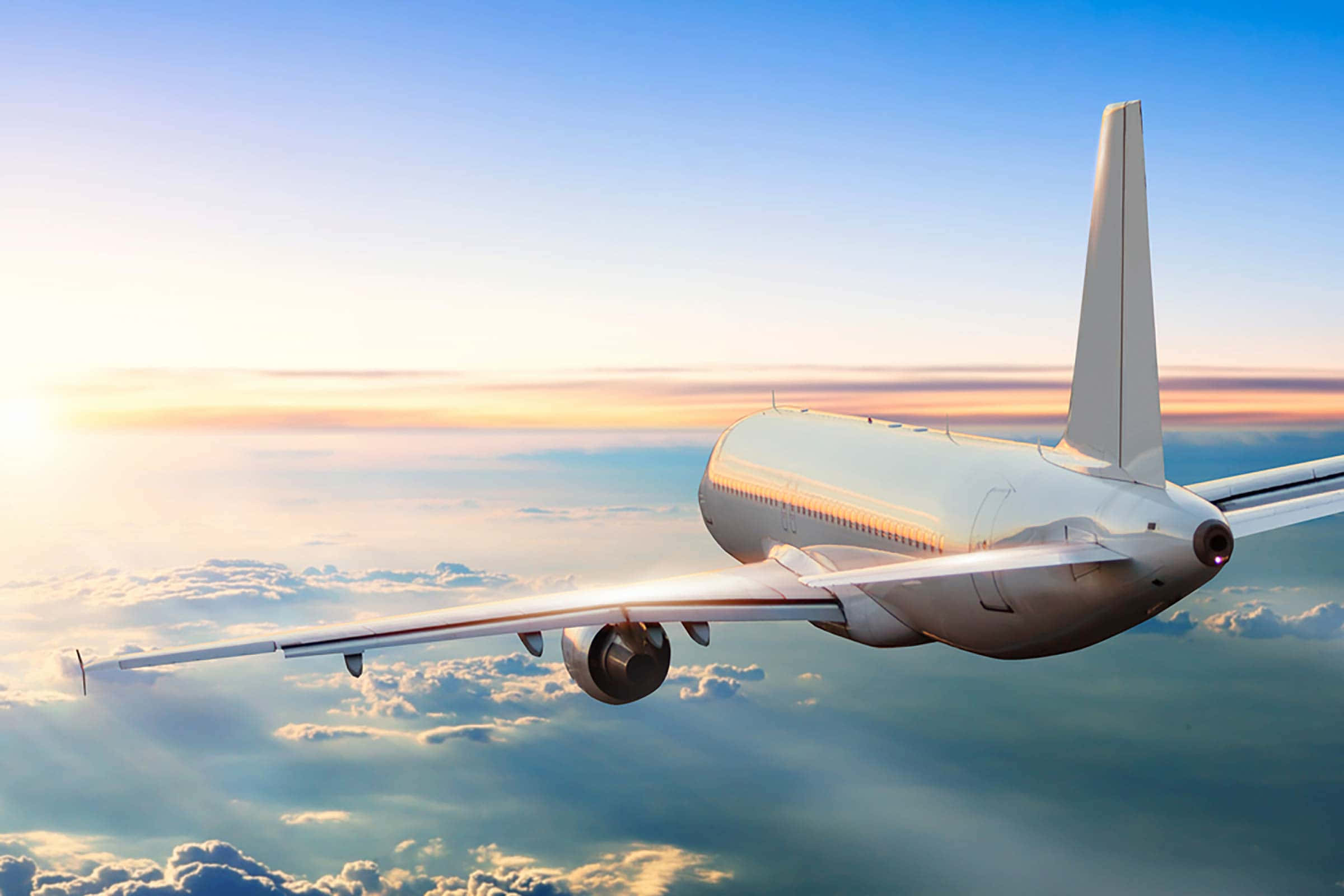 飛行飛機圖像 壁紙 — 一架流線型白色飛機在蓬鬆的雲層上方翱翔，沐浴在金色的夕陽光中，背景是漸變的藍色天空。