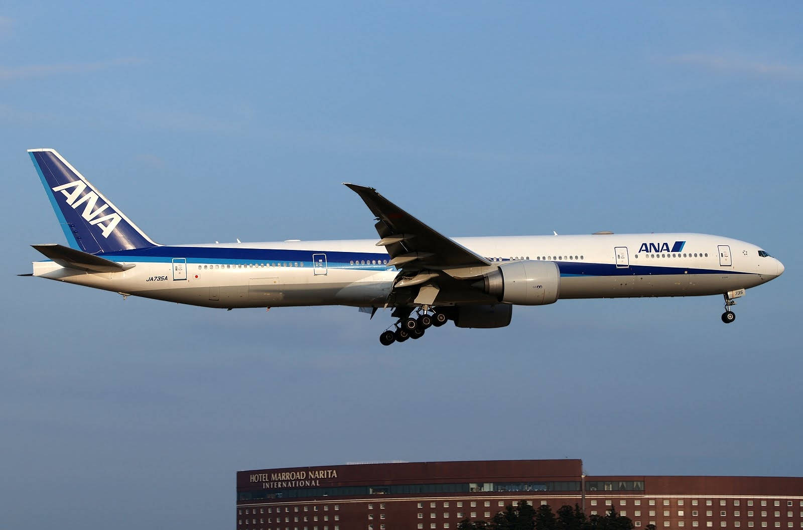 飛越成田國際機場馬羅德酒店的全日空客機 壁紙 — 一架全日空波音777在晴朗藍天下，放下起落架，降落在酒店大樓上方。