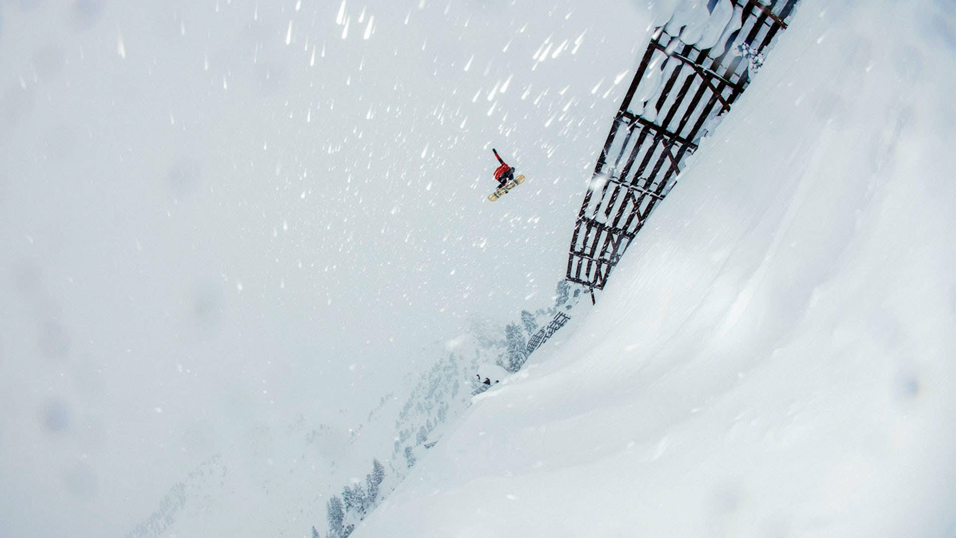飛越雪坡 壁紙 — 一名身穿紅色滑雪板運動員在陡峭的雪山斜坡上，在金屬欄杆附近飛過飄落的雪花。