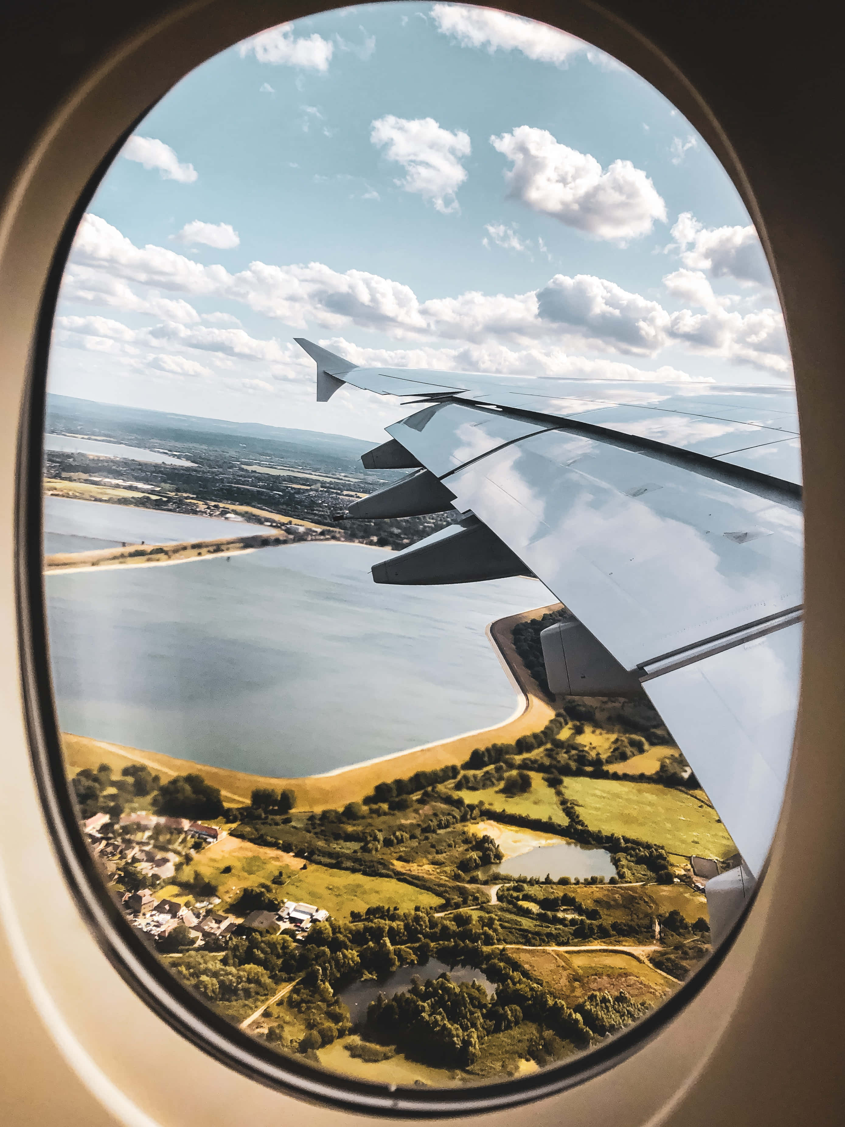 飛越雲層之上 壁紙 — 從橢圓形飛機窗戶看到的飛機機翼，俯瞰綠色田野、湖泊和散落的城鎮，天空明亮多雲。