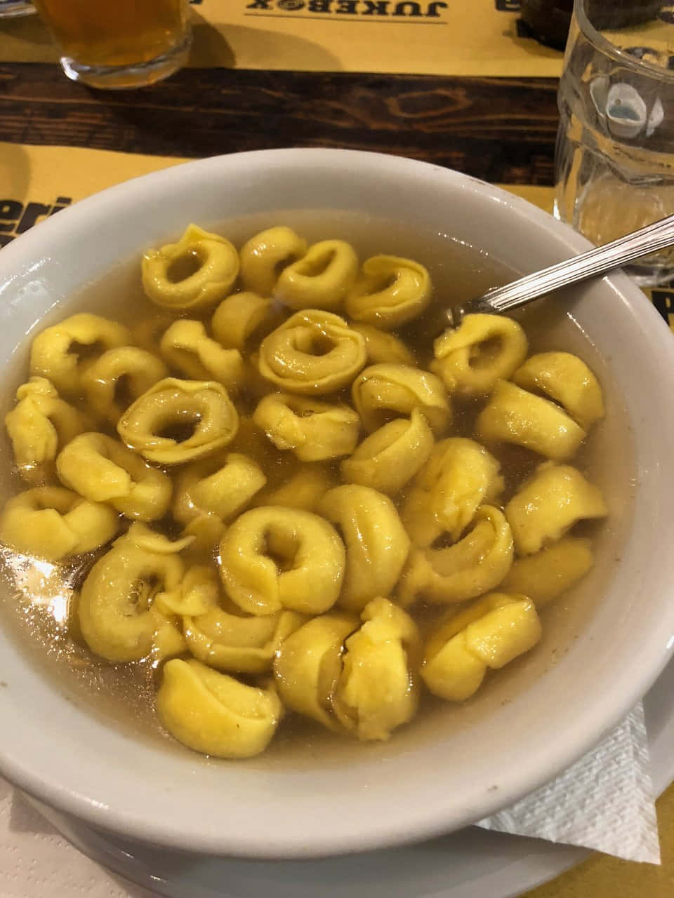餐廳中的正宗義式餃子湯 壁紙 — 一個白色碗的特寫，裡面盛滿了金黃色的意大利餃子，浸泡在清澈的湯汁中，湯匙擱在碗內。