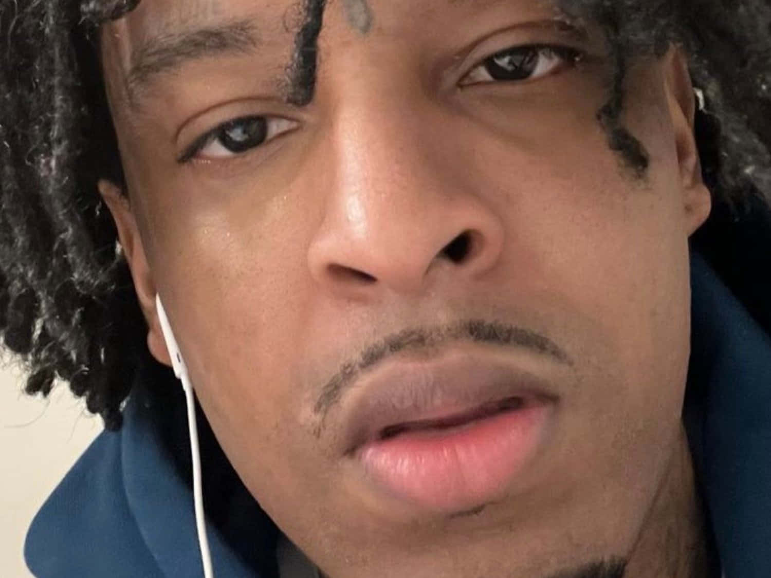 饒舌歌手 21 Savage，以獨特音色聞名 壁紙 — 一名留著捲髮、臉上有紋身的男子特寫肖像，戴著白色耳機和藍色連帽衫。