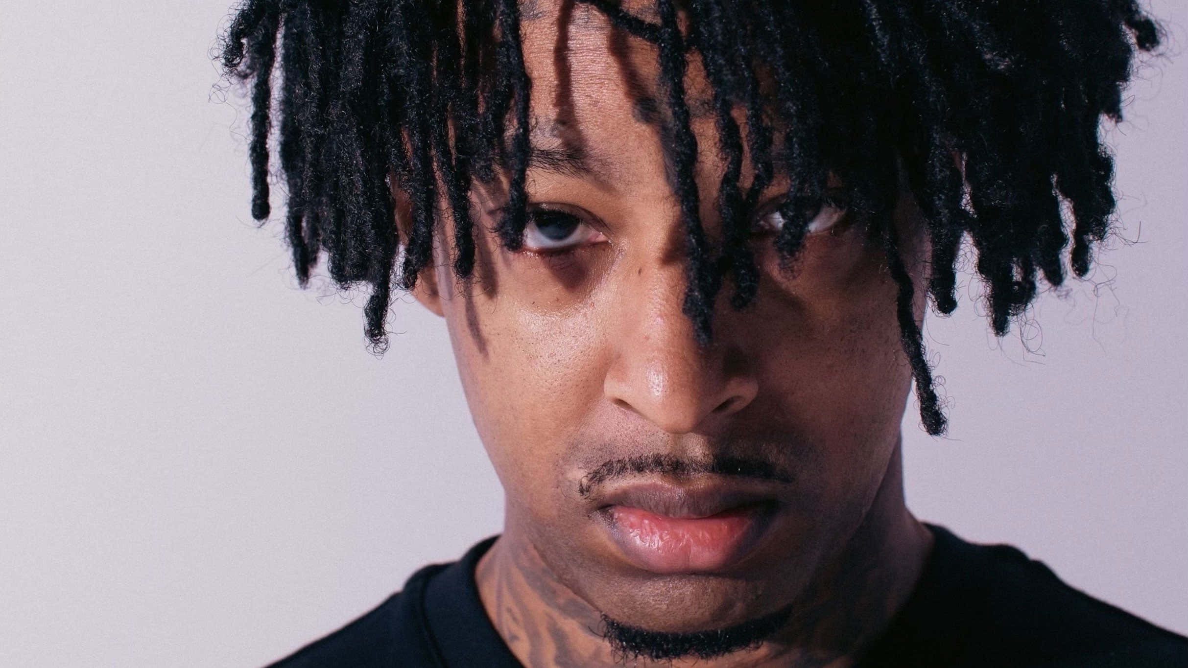 饒舌歌手 21 Savage 登台表演 壁紙 — 一名留著黑色辮子、身上有紋身的男子，在純淺灰色背景前凝視的特寫肖像。