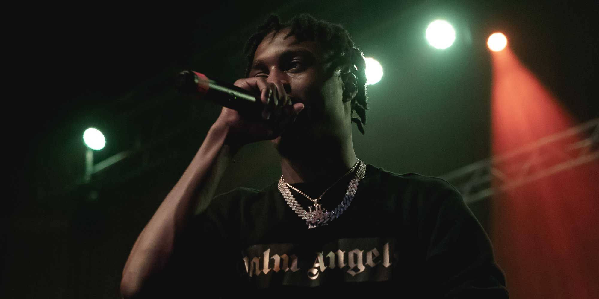 饒舌歌手 Lil Tjay 舞台演出 壁紙 — 一位說唱歌手在戲劇性的綠色和紅色聚光燈下，以強烈的專注力在舞台上表演。