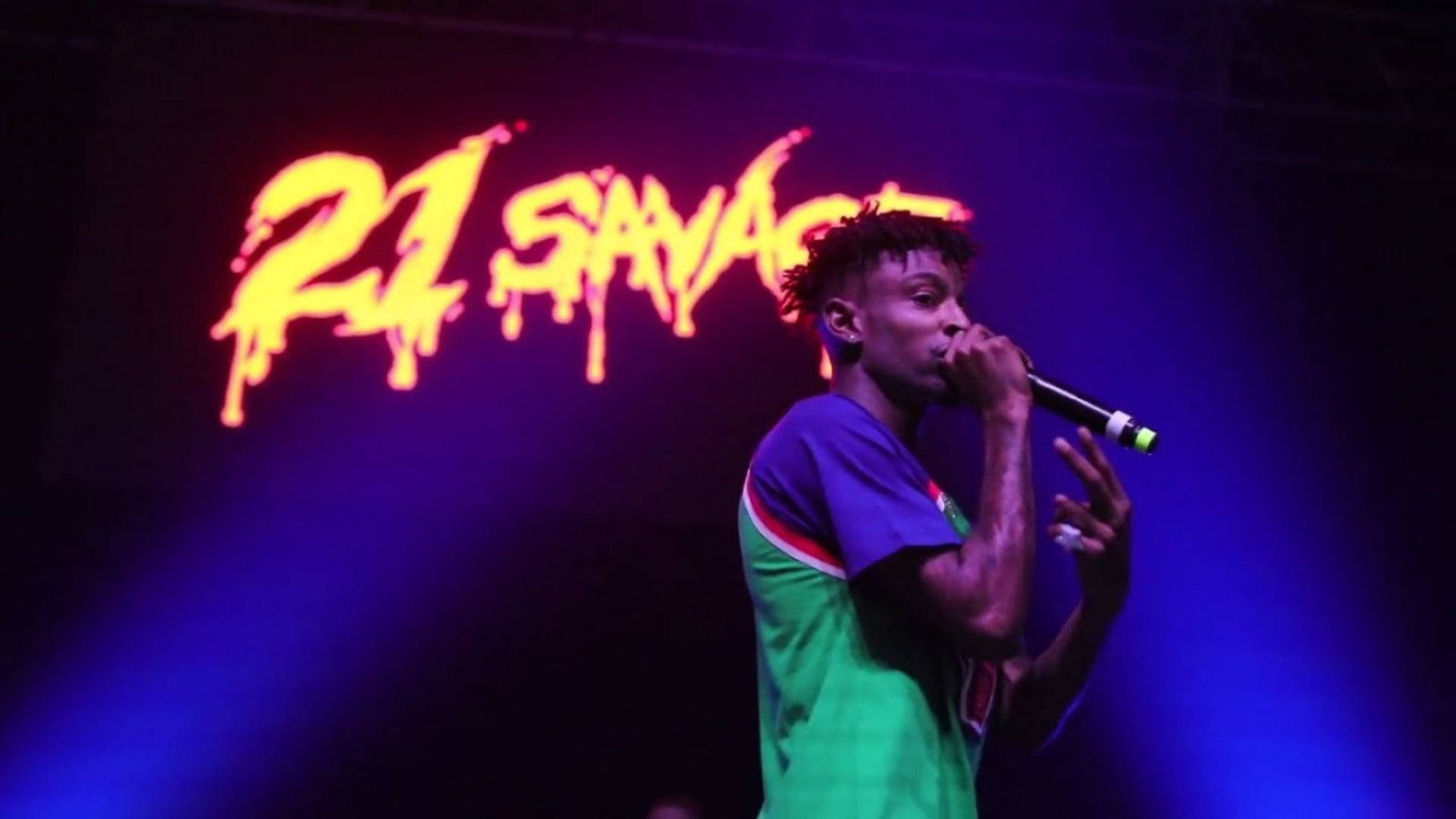 21 Savage Numb The Pain Tour 壁紙 — 饒舌歌手21 Savage在紫色燈光下於舞台上表演，他的名字在他身後以火熱的橙色發光。