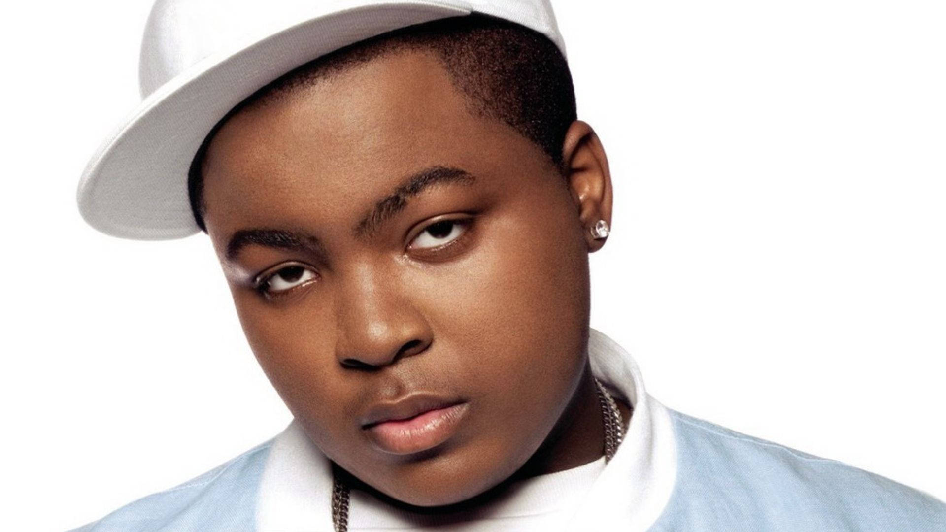 饒舌歌手Sean Kingston 壁紙 — 一位年輕黑人男子表情嚴肅，戴著白色帽子，穿著淺藍色襯衫，背景為純白色。