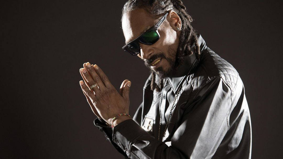 Snoop Dogg 戴著太陽眼鏡，在昏暗的攝影棚肖像中雙手合十。
