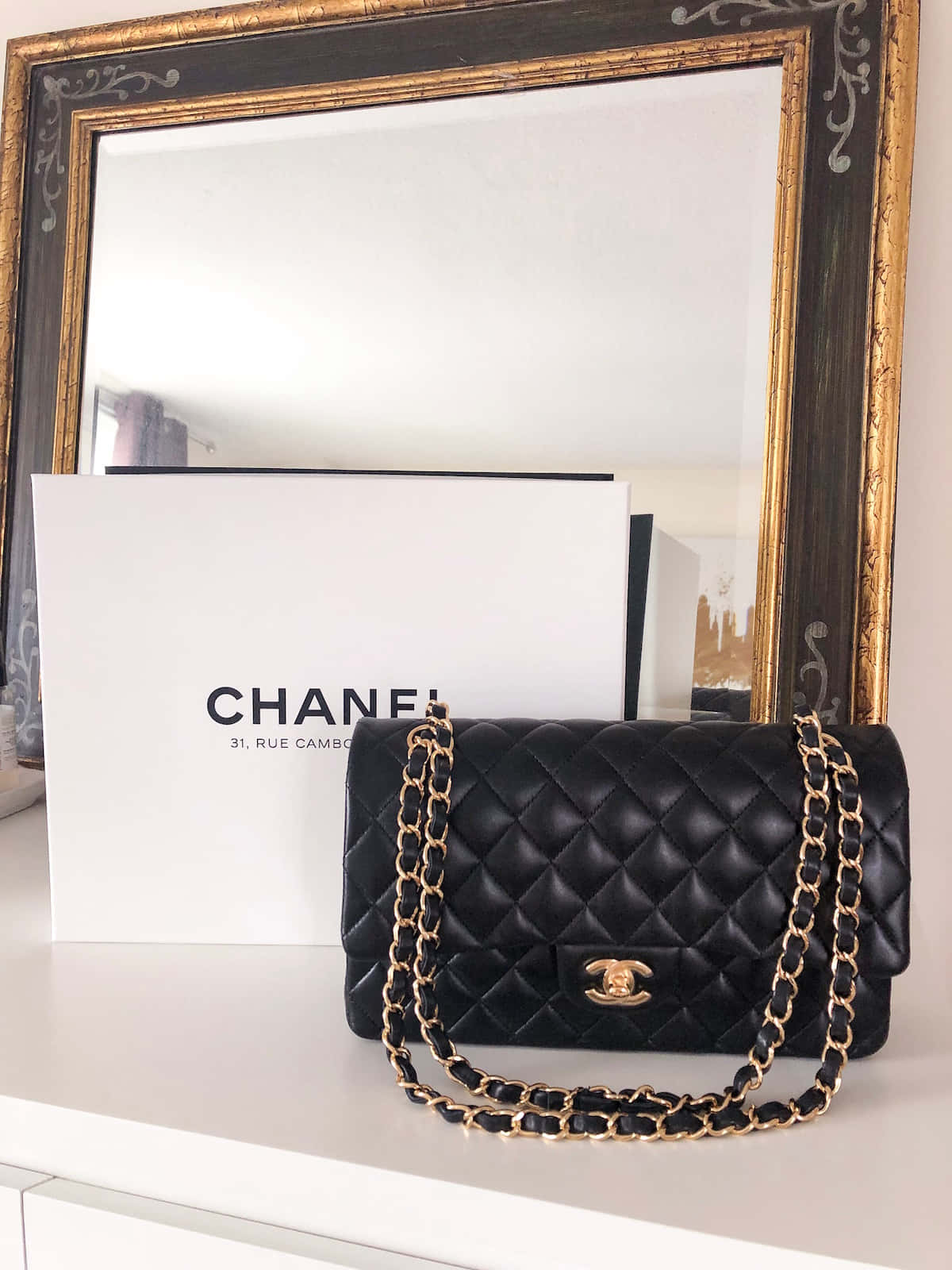 香奈兒中的自信與快樂 壁紙 — 一個黑色絎縫Chanel手袋配金色鏈條，放在白色表面上，旁邊是其品牌盒子和華麗鏡子。